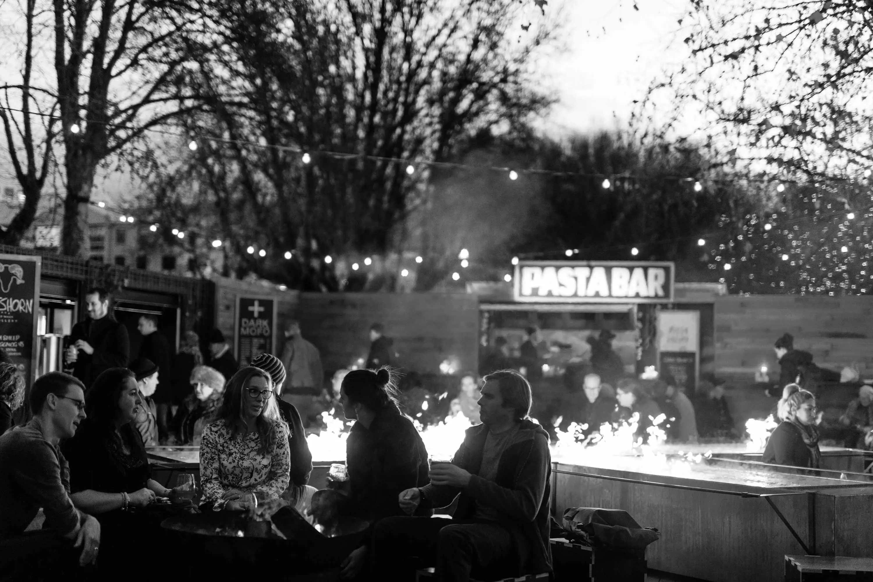 People sit outside below tree and next to large fire pits, eating, talking and drinking.