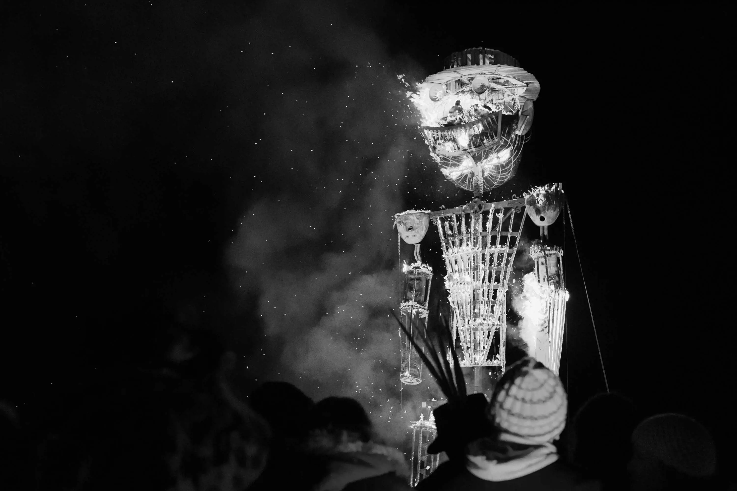 A enormous effigy of a man with a large grin and a rimmed hat made of wood and steel, burns against the night sky.