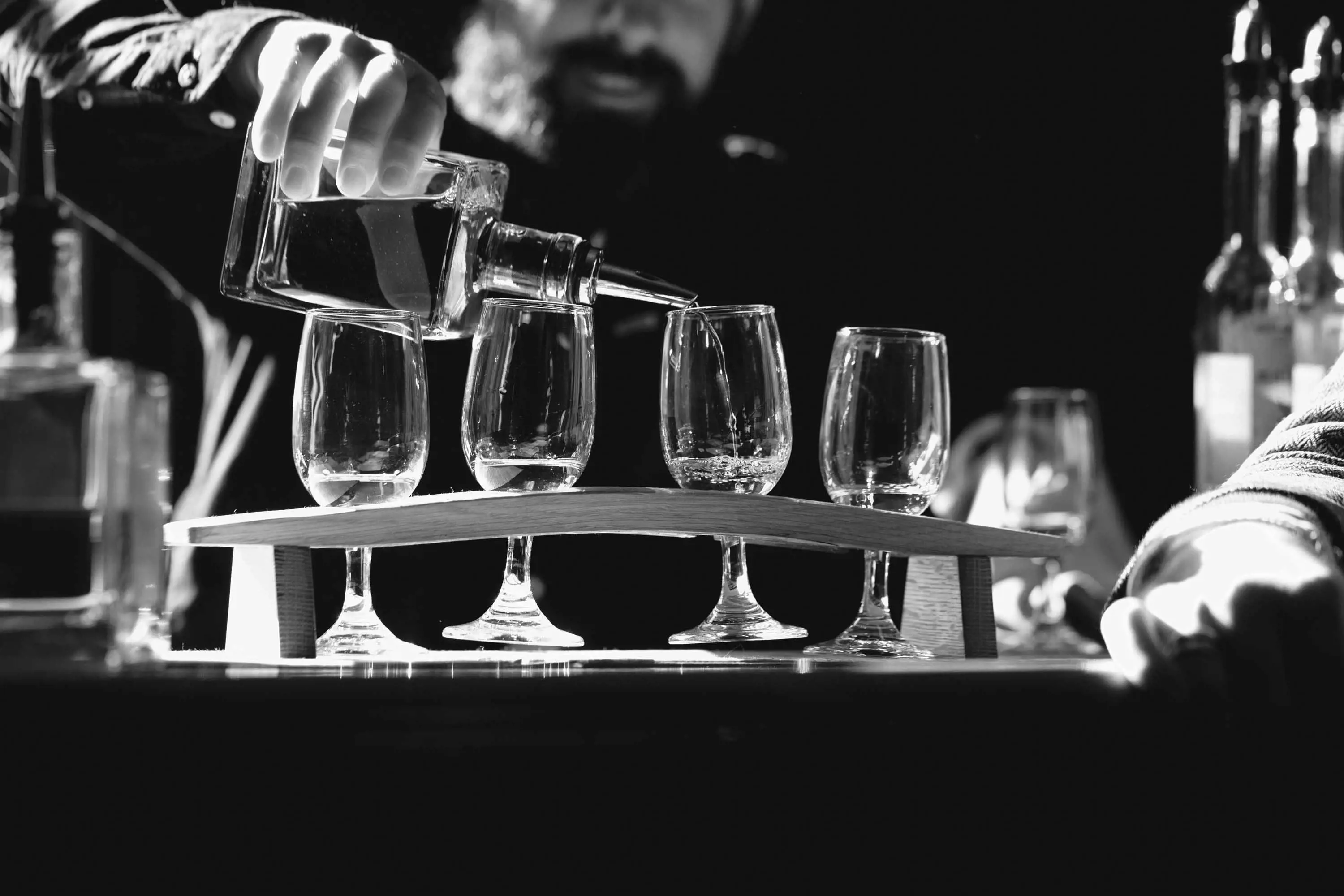 A man with a beard pours whisky from a bottle into a flight of four, tall glasses cradled in a wooden holder.