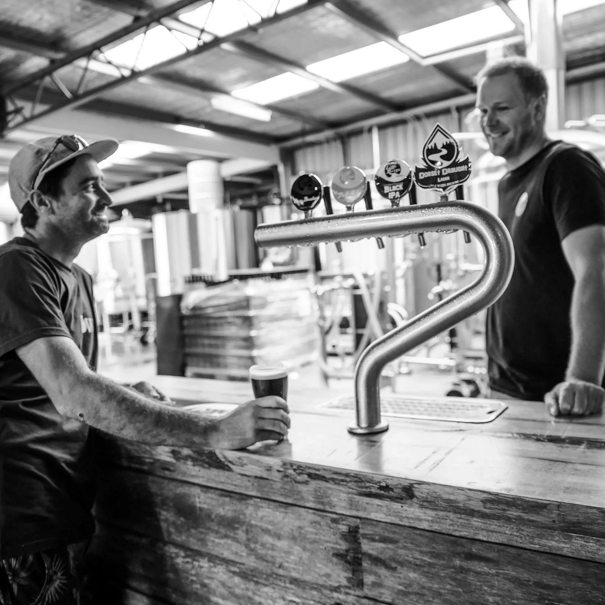 A black and white image of a man standing at a rustic wooden bar with a beer, and a bartender behind the bar, looking at a silver beer font with four taps.