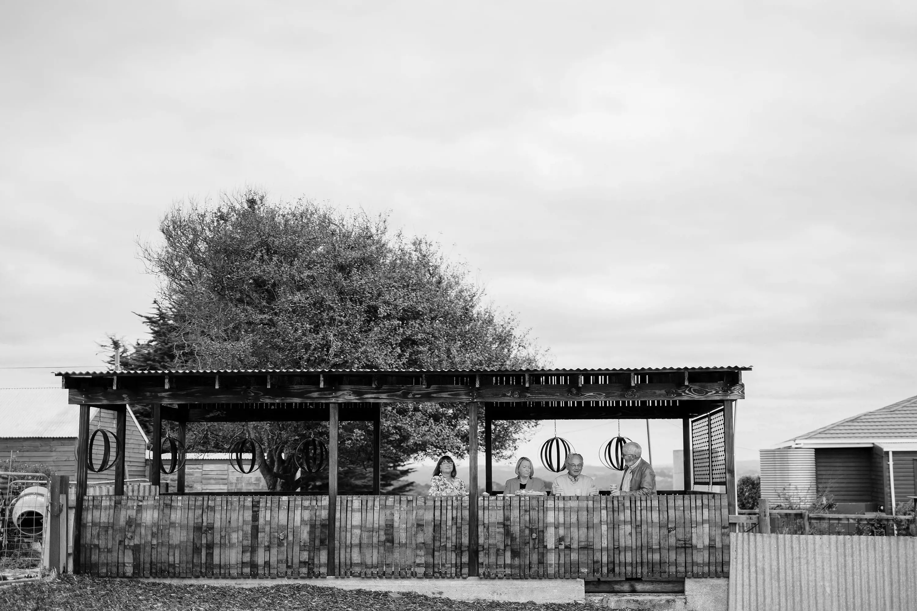 A black-and-white image of a rustic outdoor wooden shelter, with four people sitting along its bar looking over the landscape.