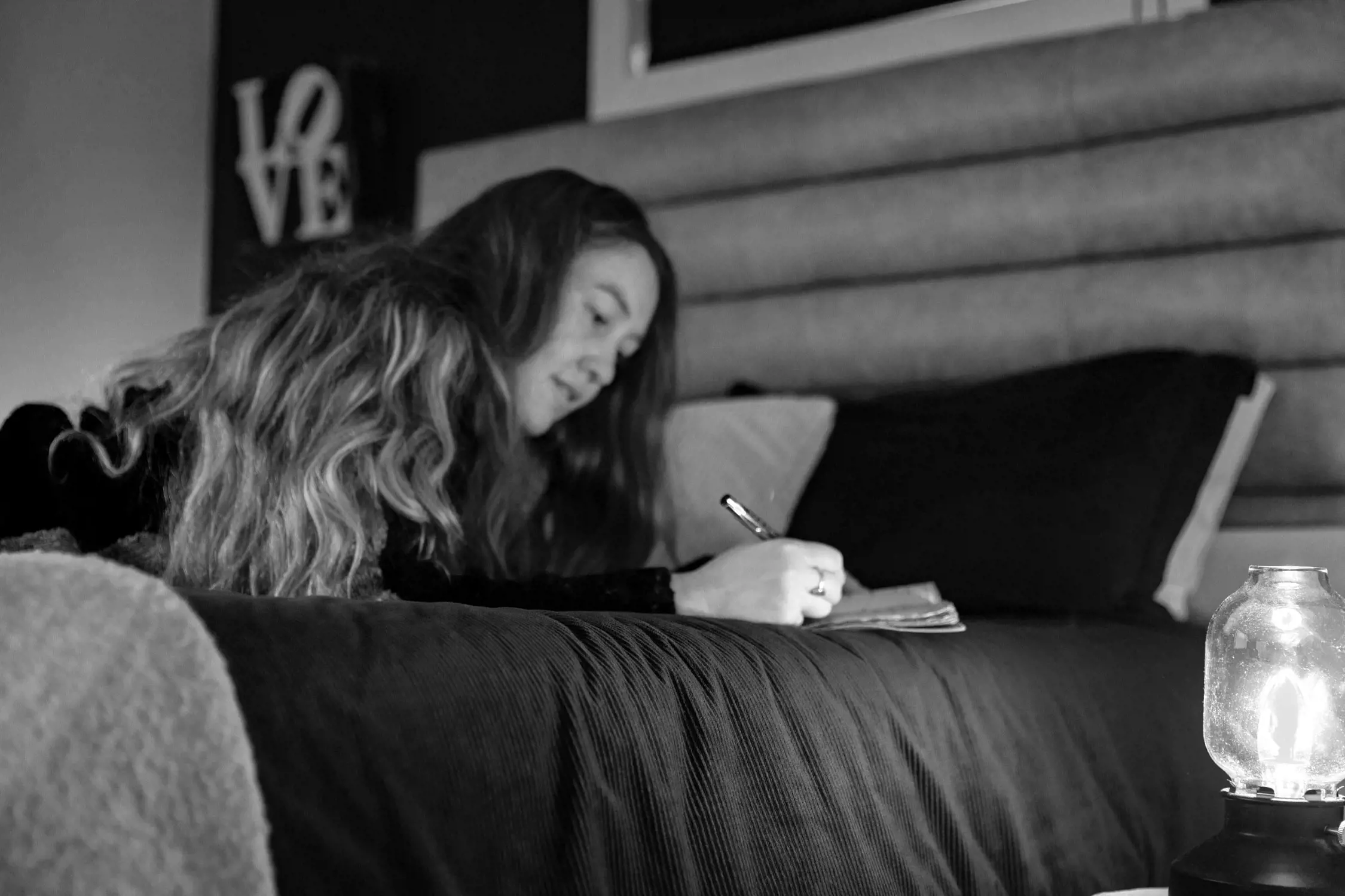 A woman with long wavy hair is lying on her stomach on a bed, deeply focused as she writes in a notebook. The bed has a dark, textured bedspread, and a warm lamp to the right illuminates the space, creating a cozy atmosphere. Behind her, a decorative sign reading 'LOVE' is partially visible on a wall.