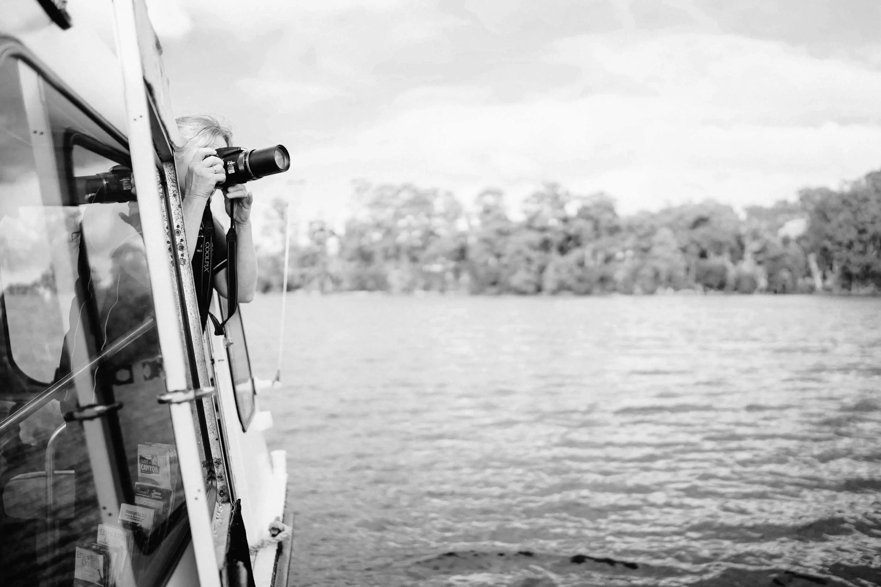 A woman with a SLR digital camera with a large zoom lens leans over a rail under the canopy of a small cruise boat and takes a photo.