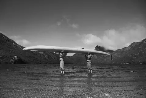 Two people hold a large canoe over their head and stand in the shallow water at the edge of a large lake in front of mountains.