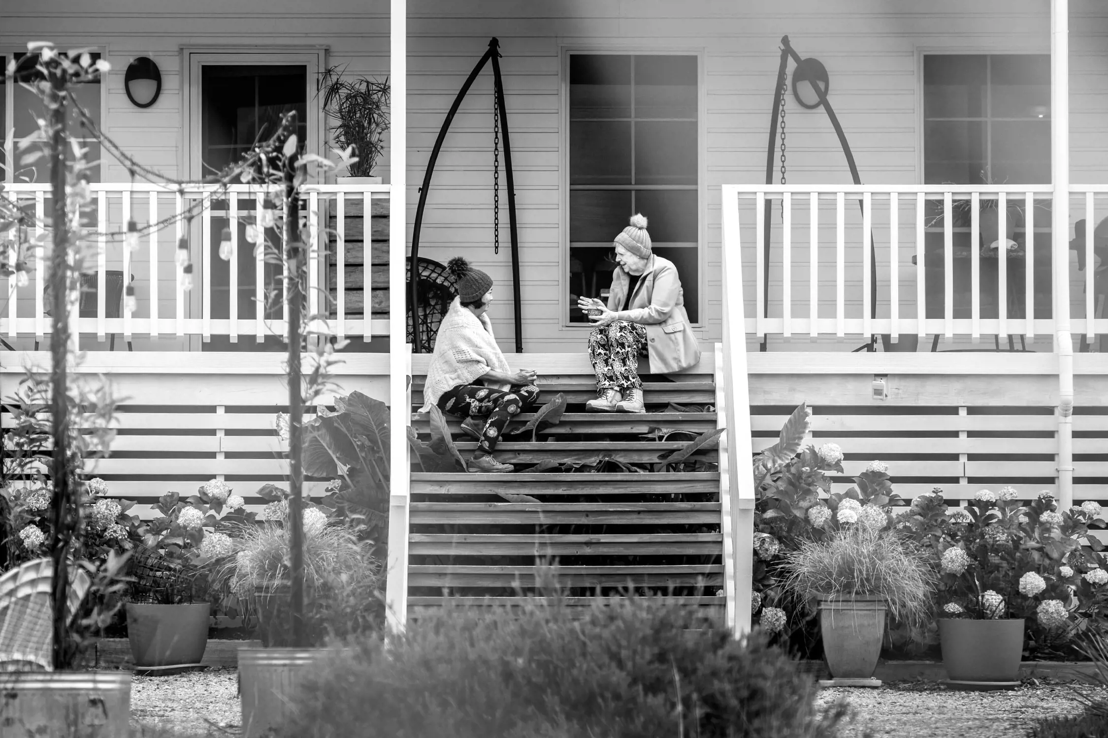 Two women are sitting on a wooden staircase leading up to a porch, engaged in conversation. They are dressed warmly, suggesting a cool day, and are surrounded by potted plants and greenery. The porch has a white railing, and the house behind them has a quaint, inviting feel.