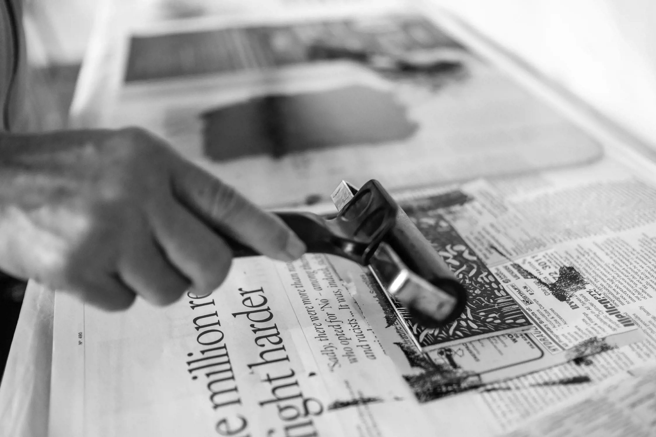 A close-up of a hand using a brayer roller to evenly spread thick black ink onto a linocut print placed on top of a sheet of newspaper. The scene captures the intricate details of the printing process, emphasizing creativity and craftsmanship