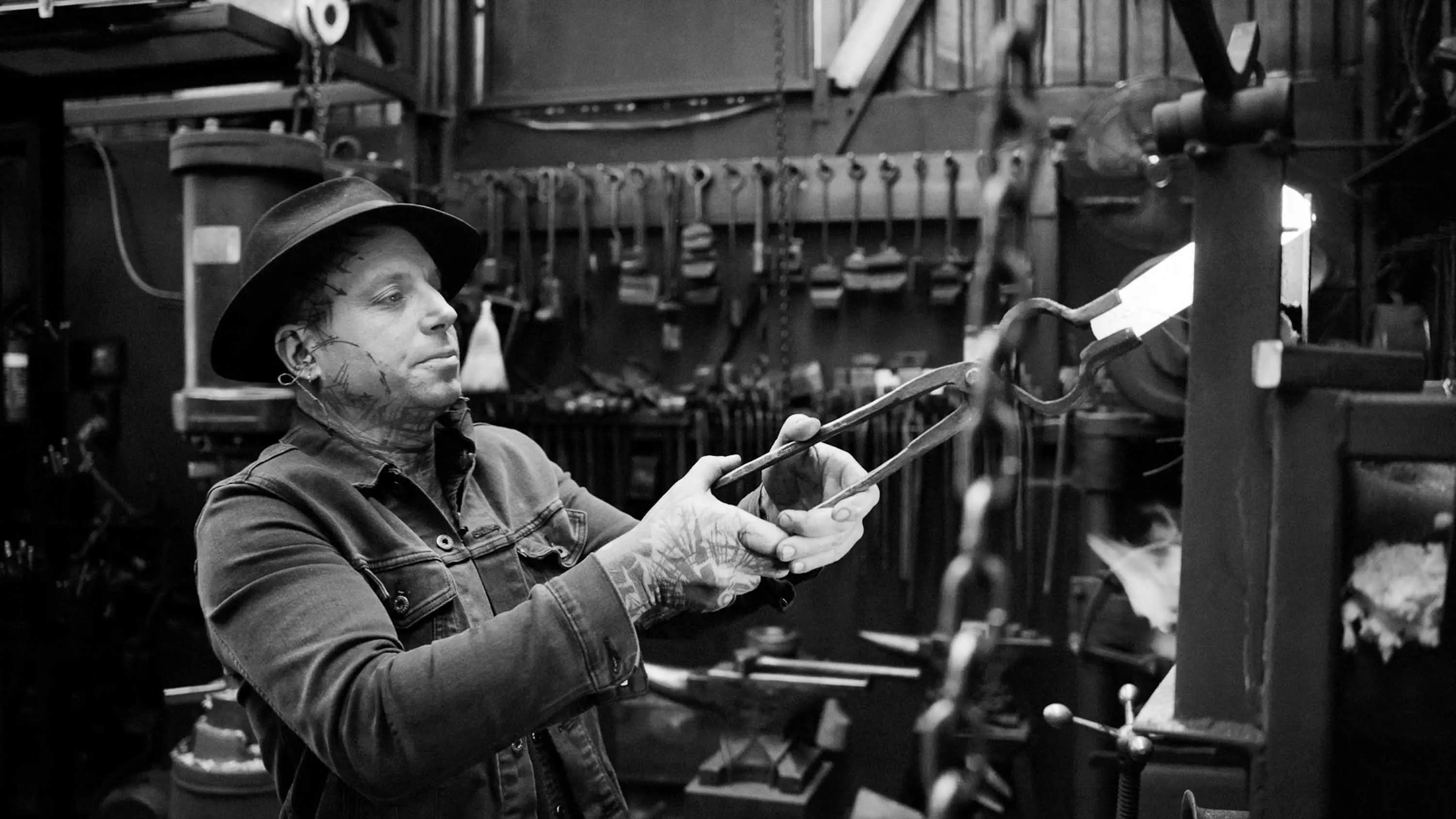 A man with tattoos wearing a hat and denim jacket holds white-hot metal in a pair of pincers in a metal workshop. 