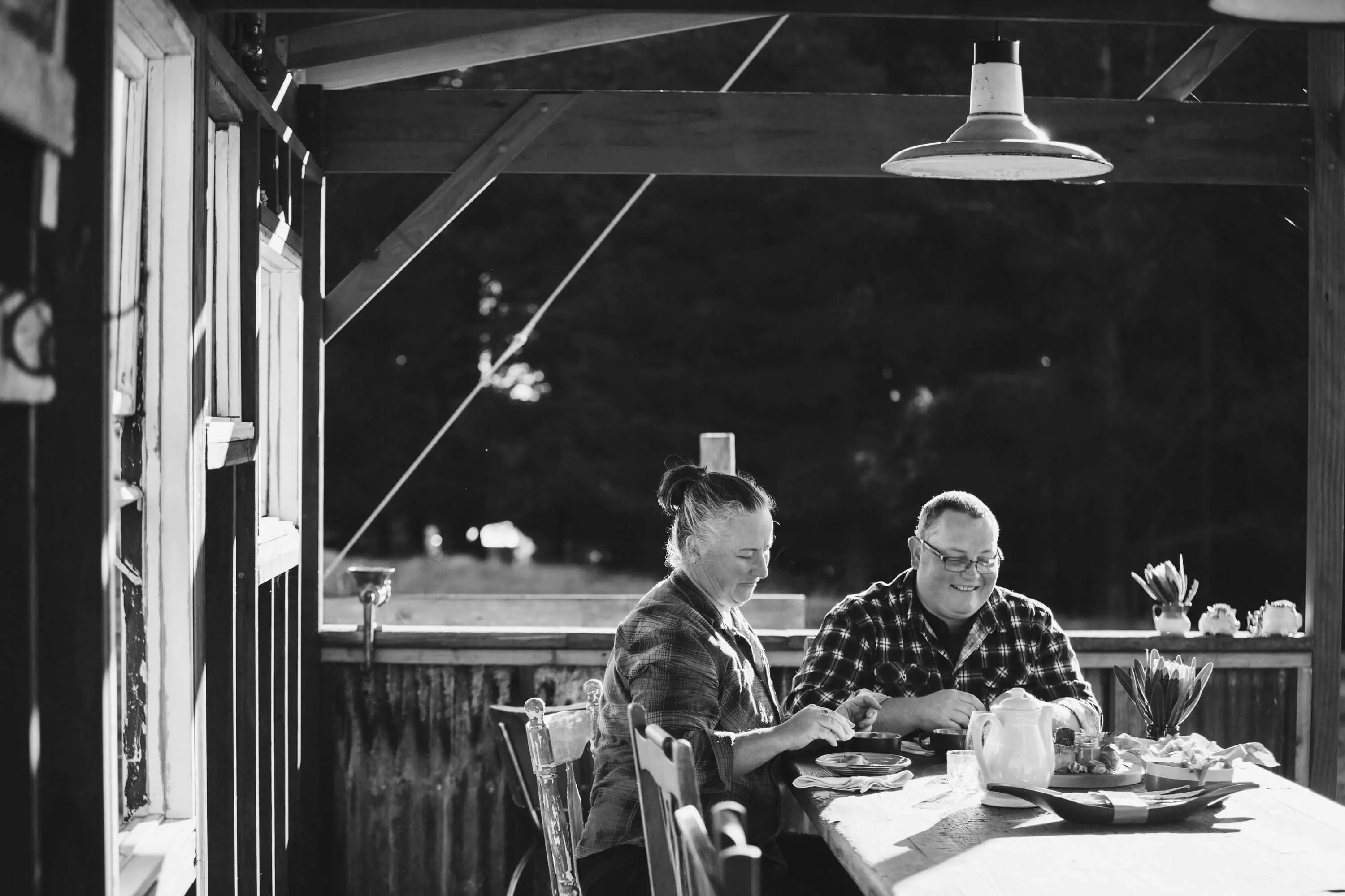 A couple sit at a large wooden table in a sheltered area outside.