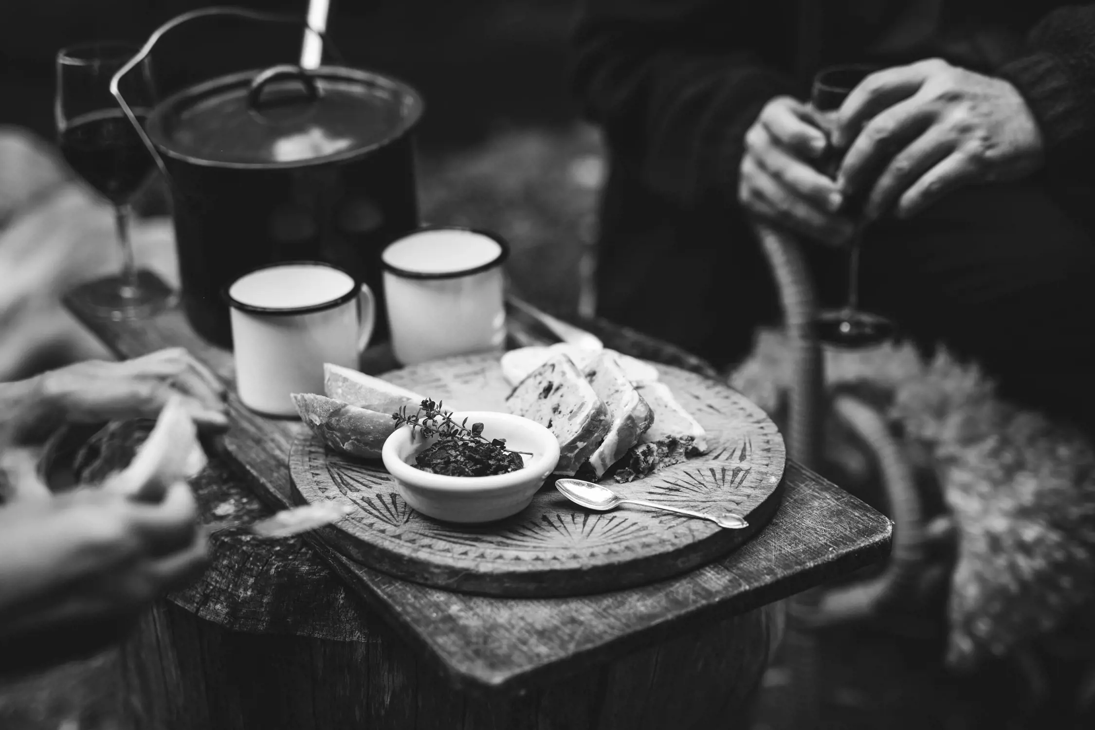 A close-up view of a rustic dining setup outdoors. A round wooden serving board holds slices of bread, a small dish of olive tapenade garnished with herbs, and other rustic food items. Two enamel mugs and a soup pot sit nearby, with blurred hands holding wine glasses in the background, indicating a cozy, shared meal