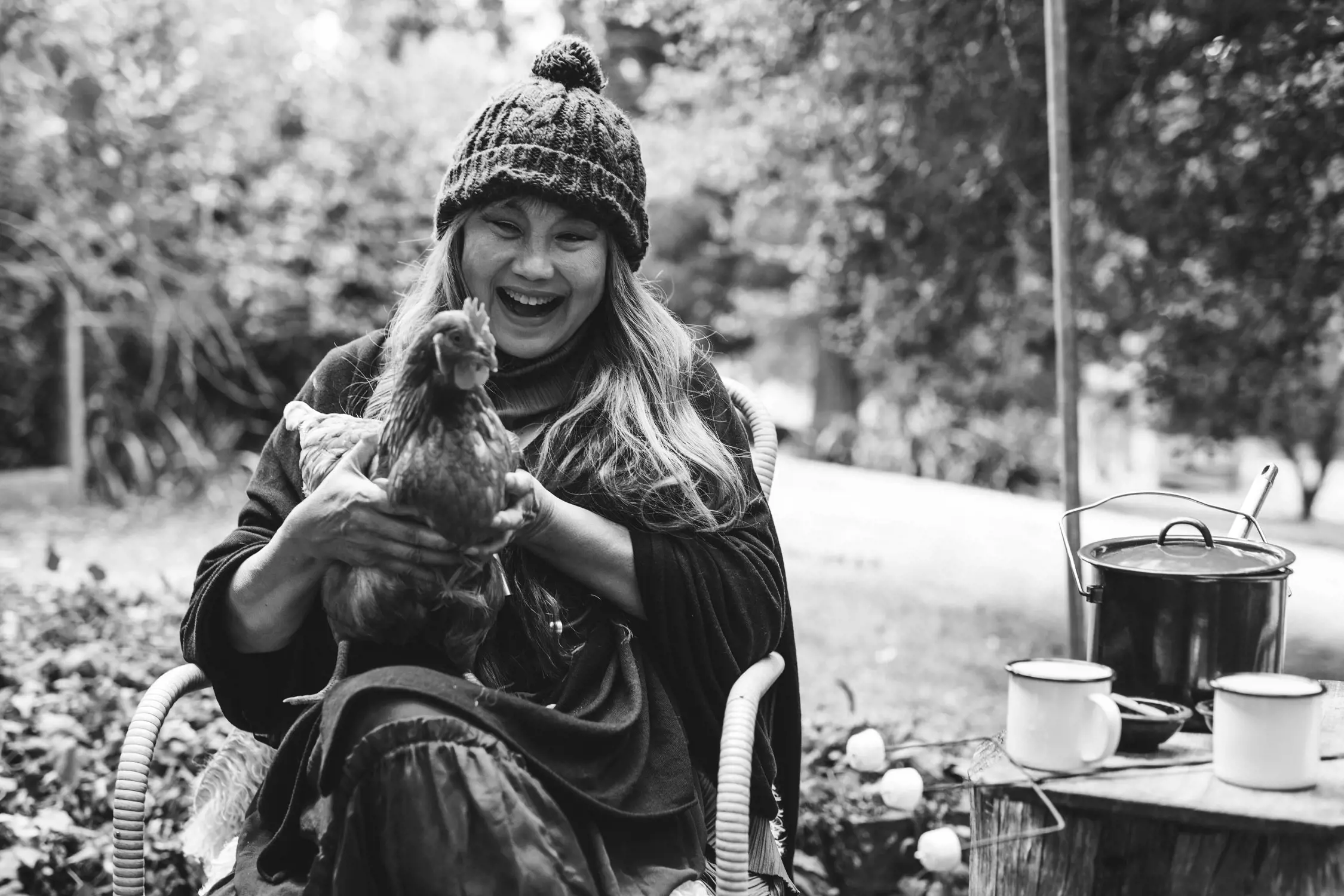 A woman seated outdoors laughs joyfully while holding a hen close to her chest. She wears a knit hat and cozy layers. Nearby, a rustic table holds enamel mugs and a cooking pot, adding to the lively, warm atmosphere of the scene