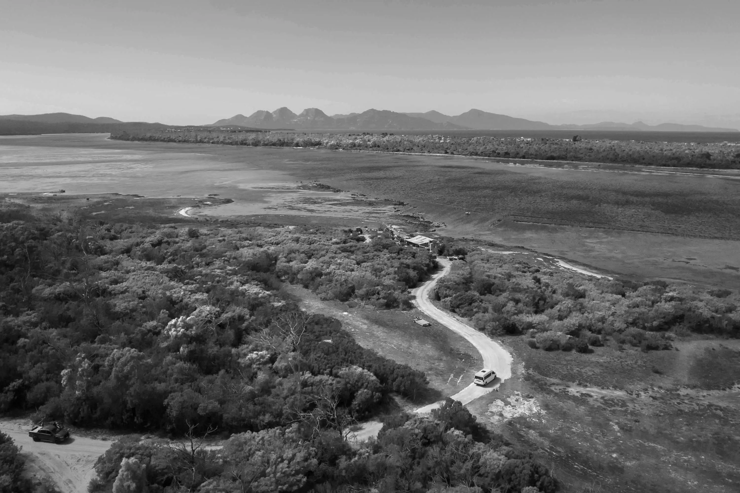 A road winds through thick scrubby bushland towards a small hut near a body of shallow water. Mountains are in the far distance.