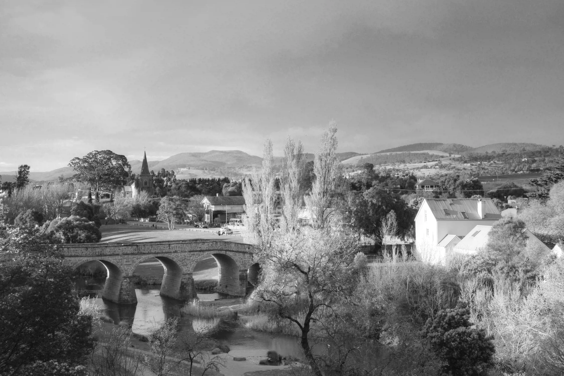 A historic stone bridge arches over a calm river in Richmond, Tasmania. Nearby, charming cottages and tall trees line the area, with a distant church steeple rising against a backdrop of rolling green hills under a peaceful sky