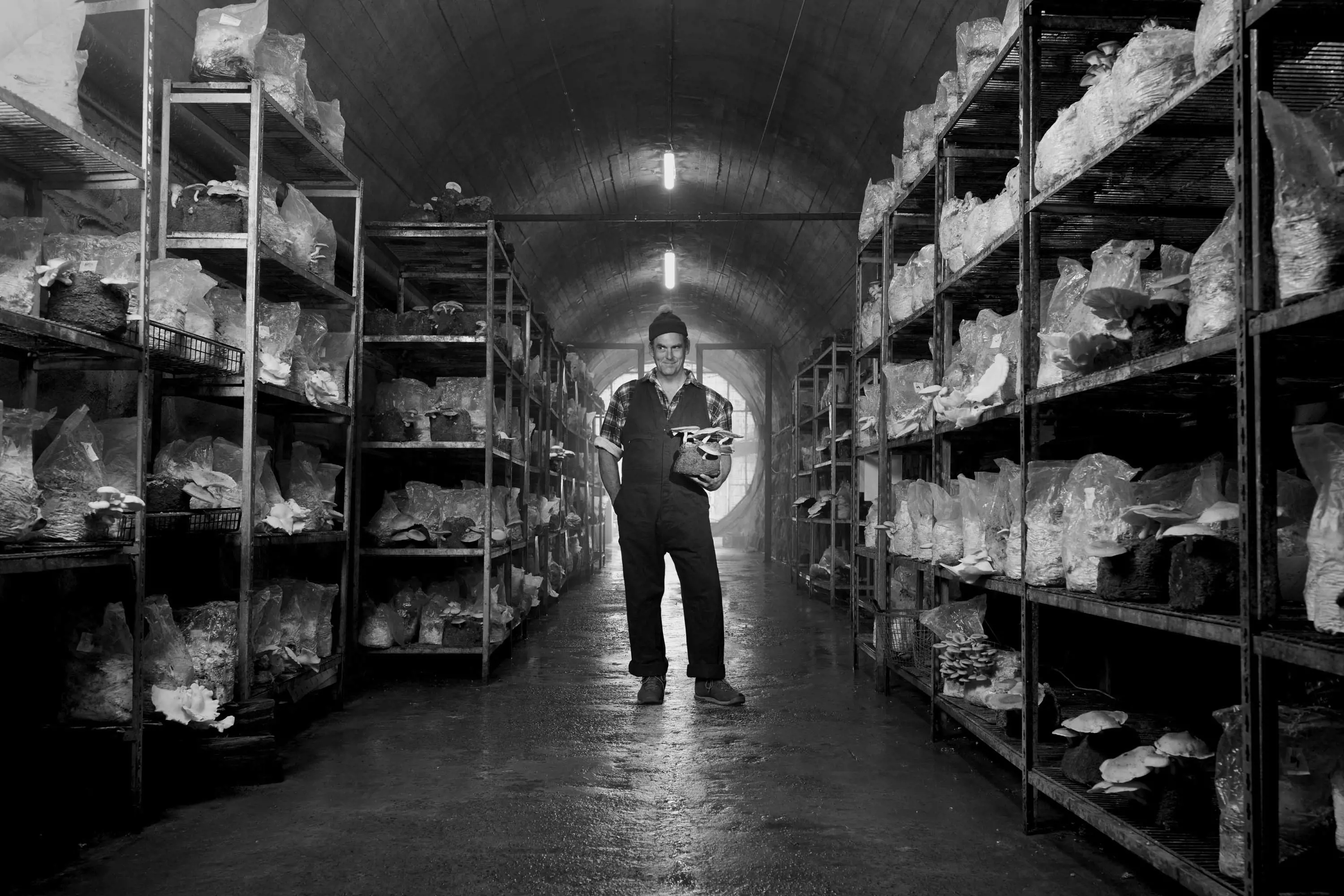 A man holds a mushroom and stands in the middle of a long, dark tunnel.