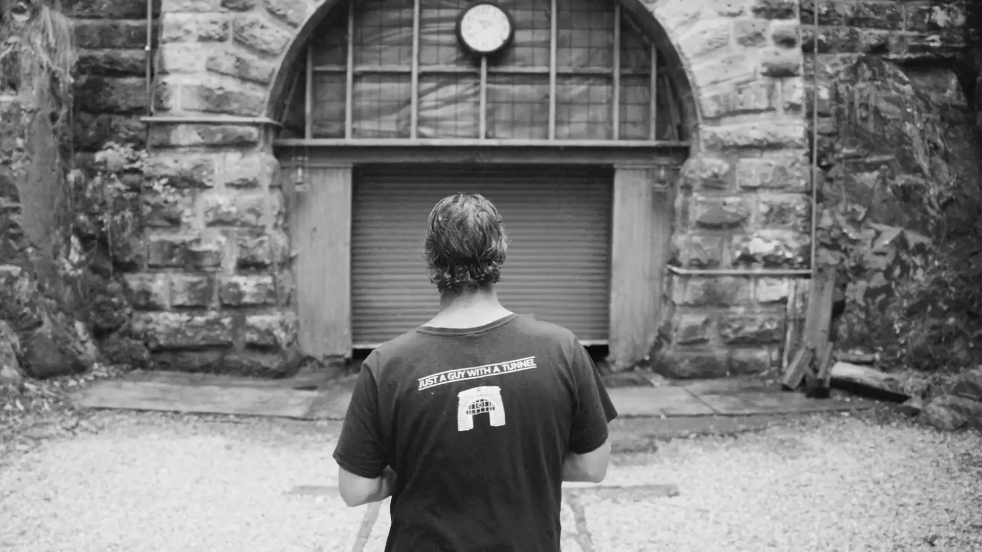 A man wearing a dark t-shirt that reads 'just a guy with a tunnel' on the back, walks towards a tunnel entrance. 