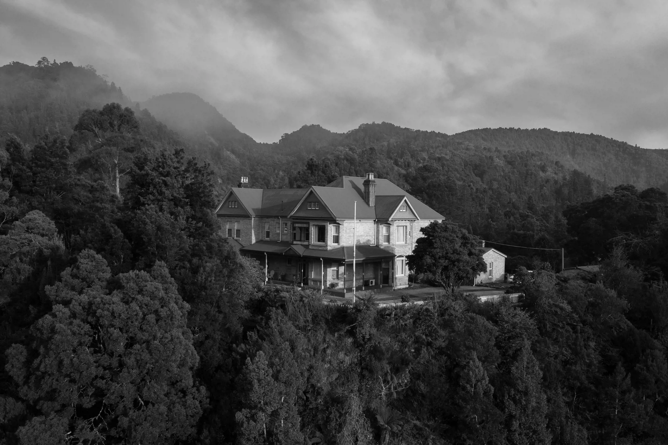 An aerial view of a grand historic building with steep roofs, situated on a forested hillside. The surrounding trees are dense and mist rises gently, creating a tranquil and scenic atmosphere.