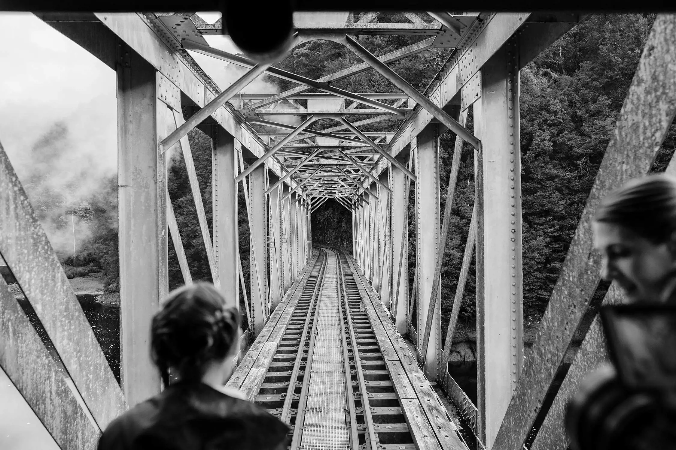 From the back of a moving train, the silhouettes of two people are looking out onto the metal and timber structures of a heritage railway bridge disappearing behind them.