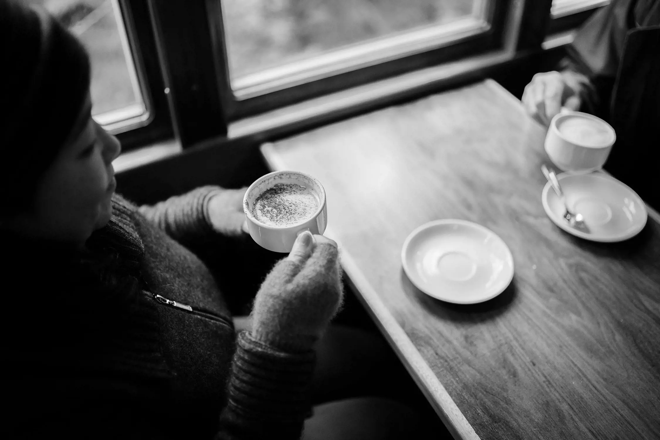 Two people sit at a wooden table inside a train carriage, holding cups and saucers with warming hot drinks. One person has cozy-looking fingerless gloves on her hands as she cradles the cup.