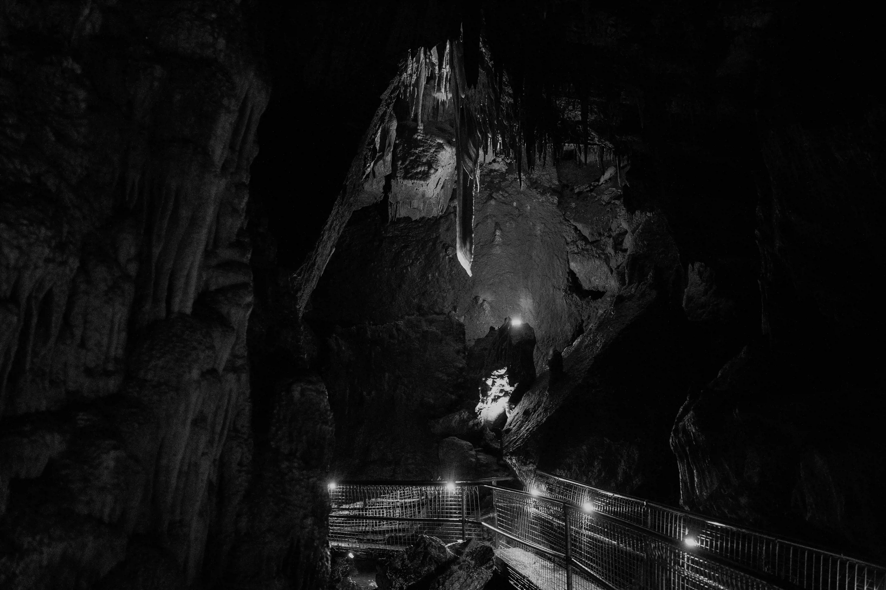 A large cave with a single stalactite suspended from the roof above a well-lit pathway.
