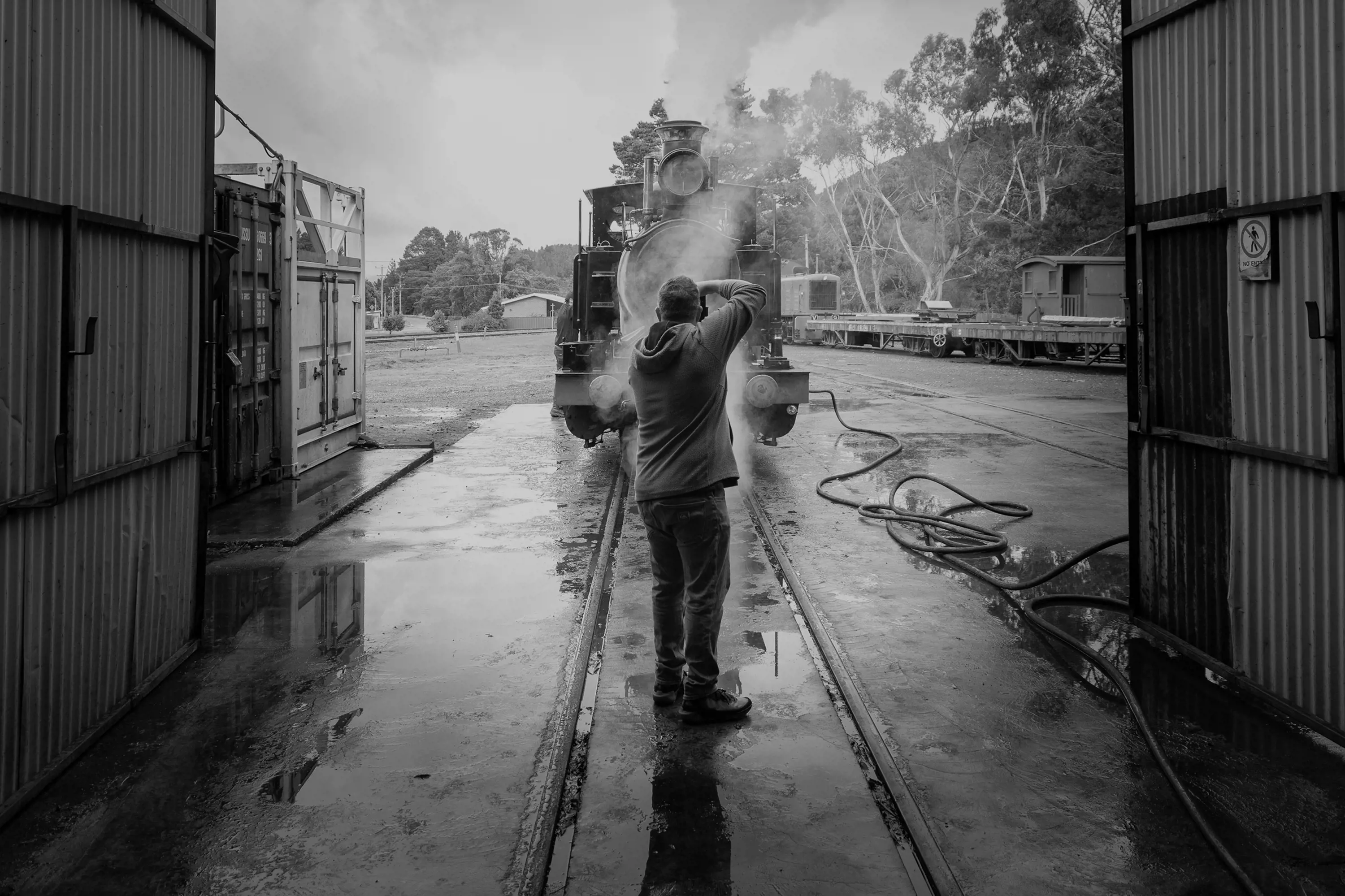 A man stands on the tracks of a locomotive steam train in a maintenance yard and takes a photograph.