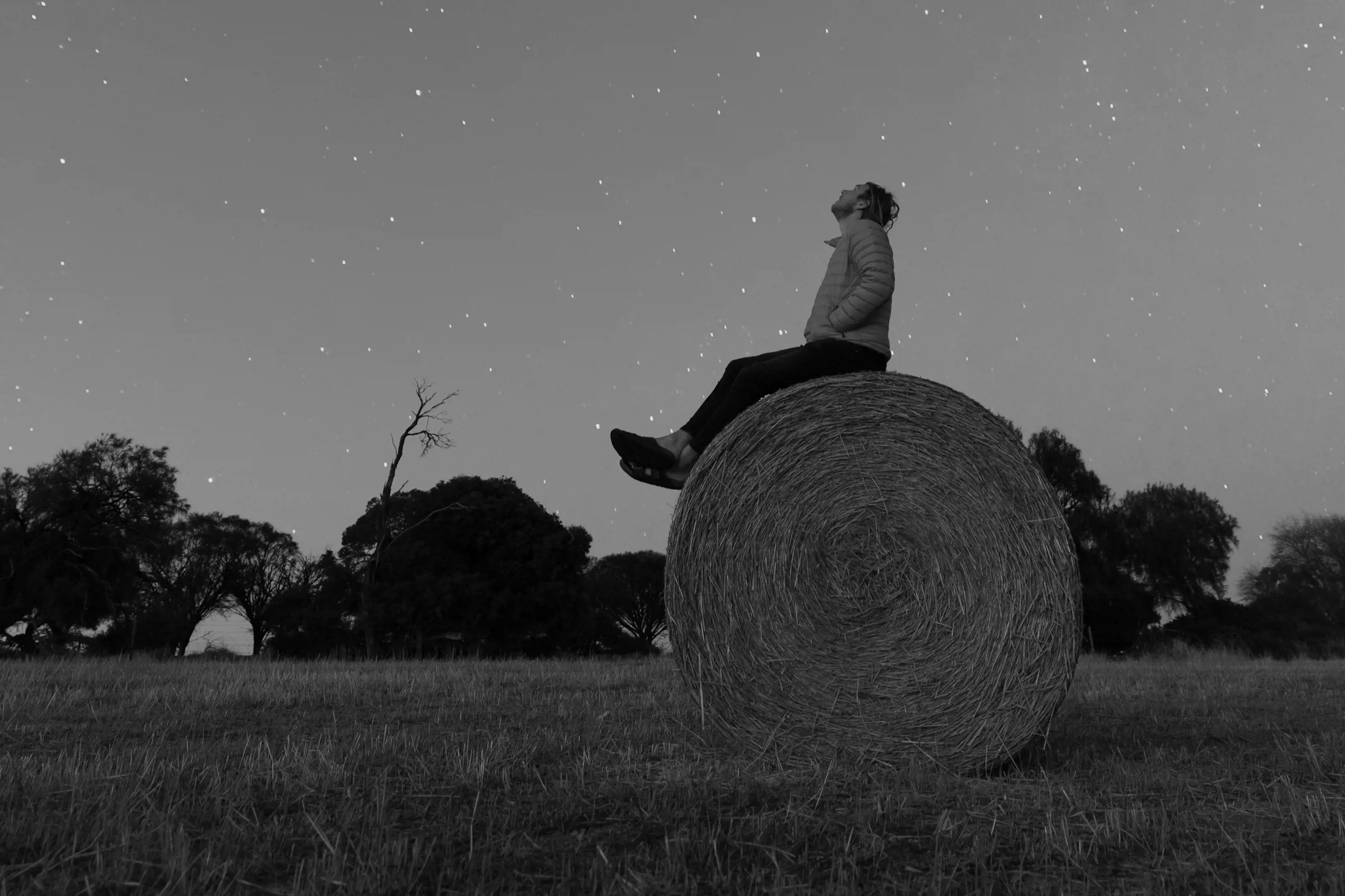 A man wearing a winter jacket sits on the top of a large cylindrical-shaped hay bale and looks up to the stars on a clear night.