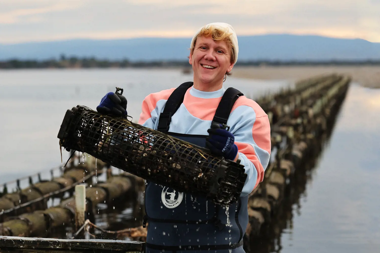 A man stands in water next to a line of oyster nest. He is wearing waterproof- waders and a beany.