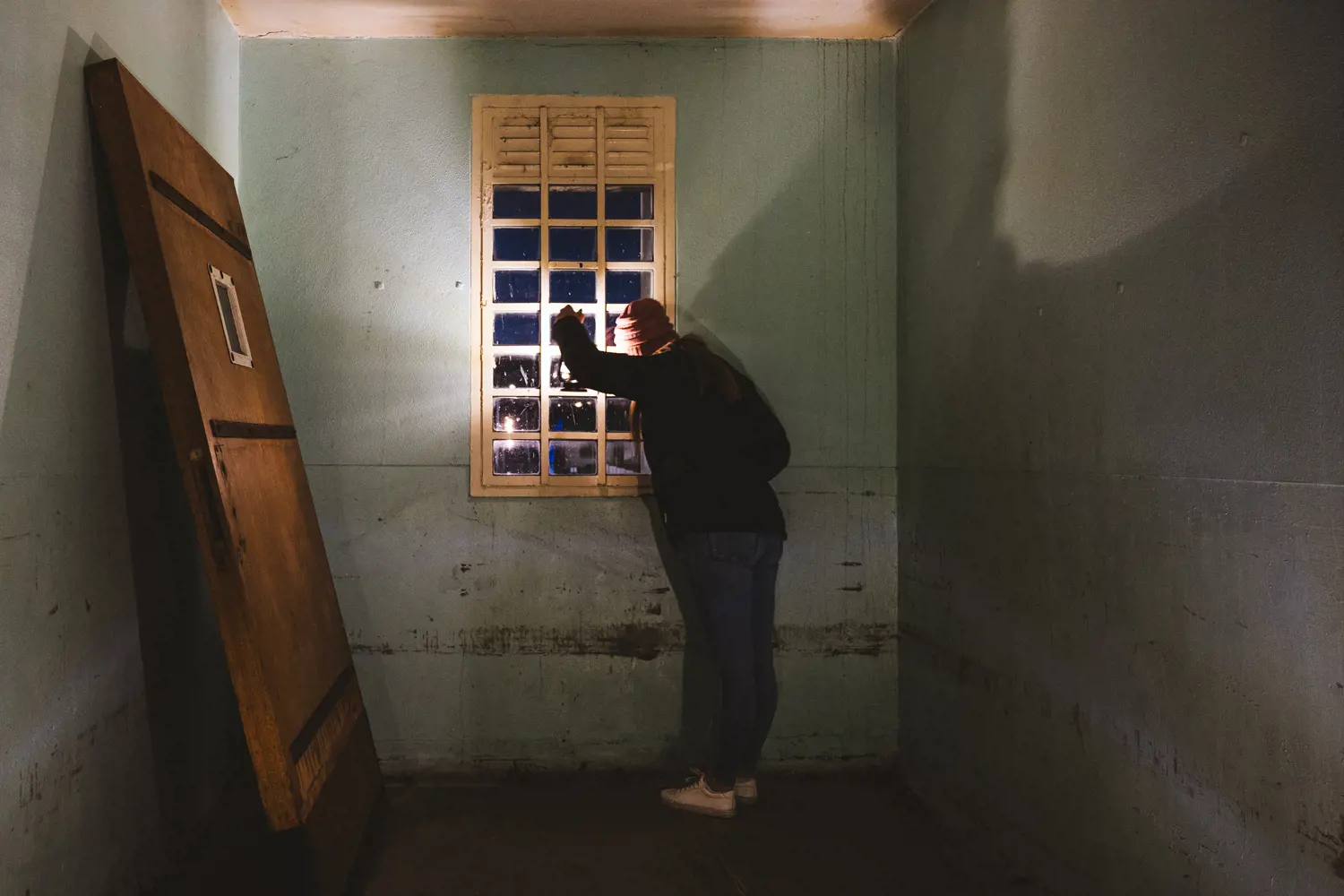 A woman wearing a beanie holds a lantern to a window at the end of a corridor in a colonial building. An old, unhinged door is propped up against a wall beside her.