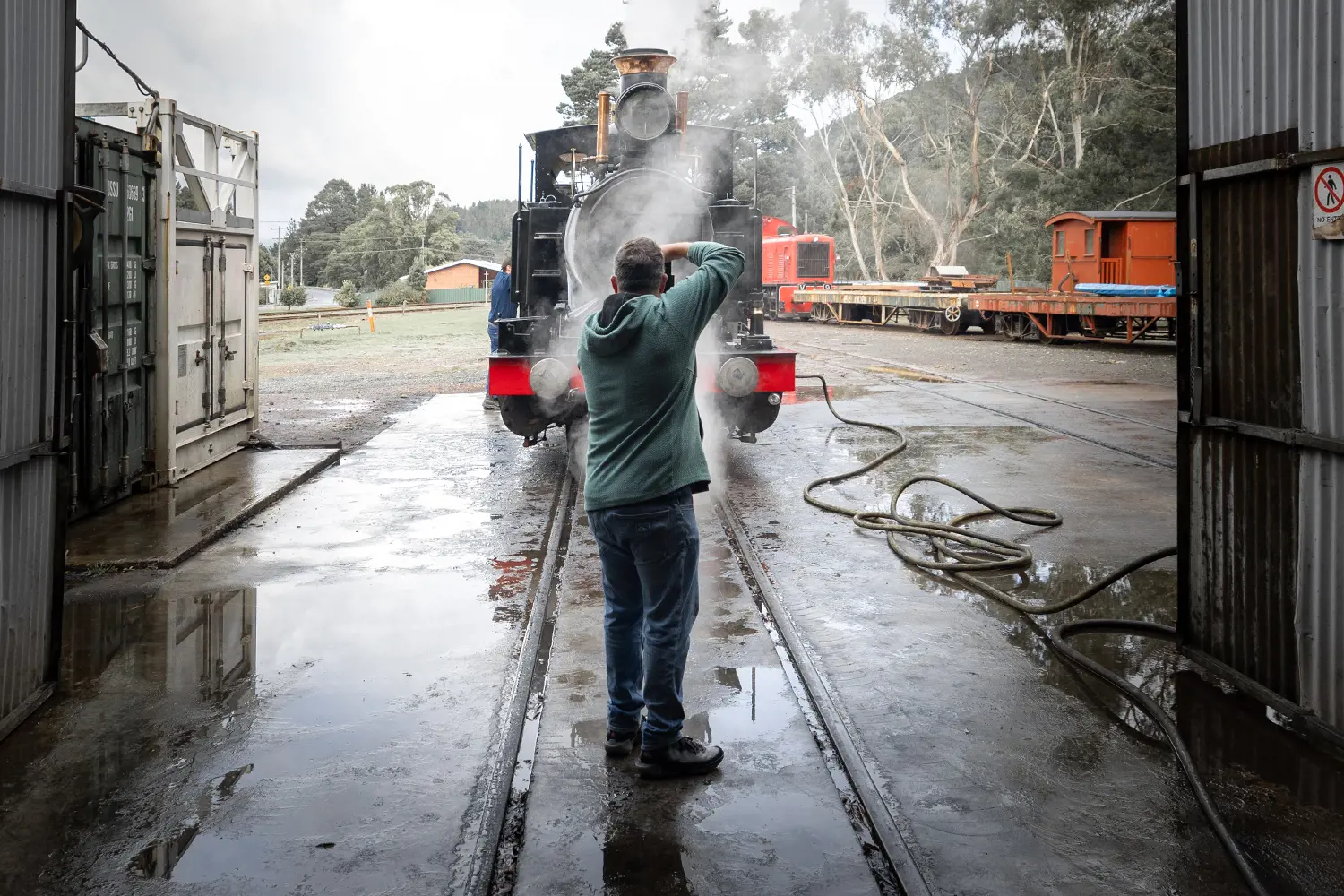 A man stands on the tracks of a locomotive steam train in a maintenance yard and takes a photograph.