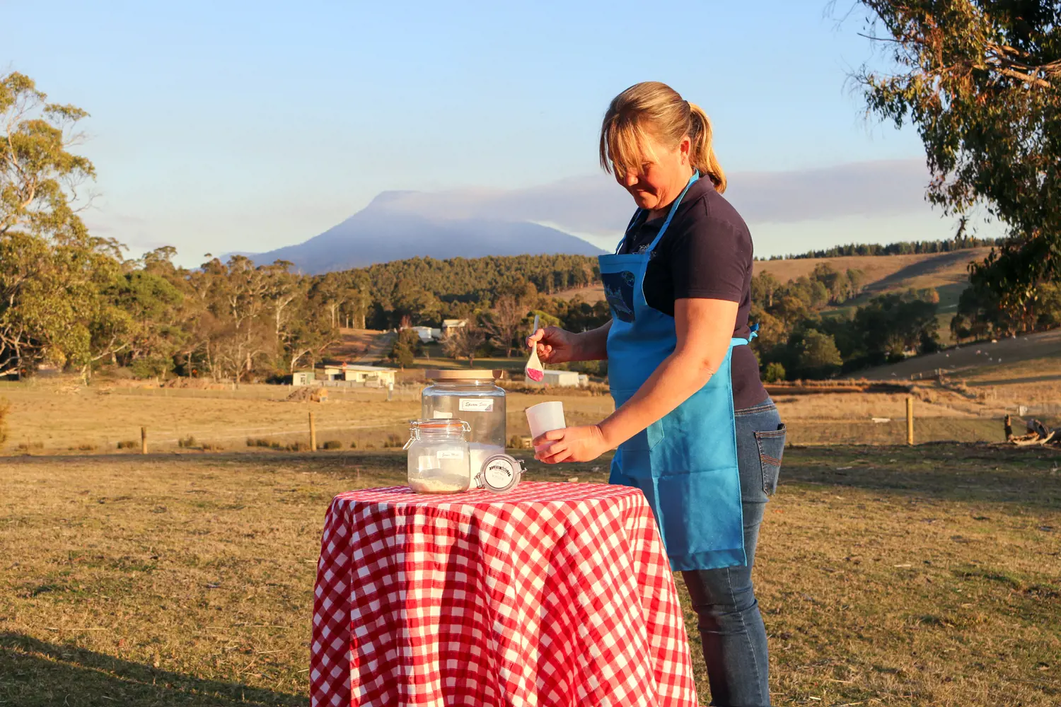 A woman wearing an apron stands in a farm field next to a table with the ingredients for making soap.