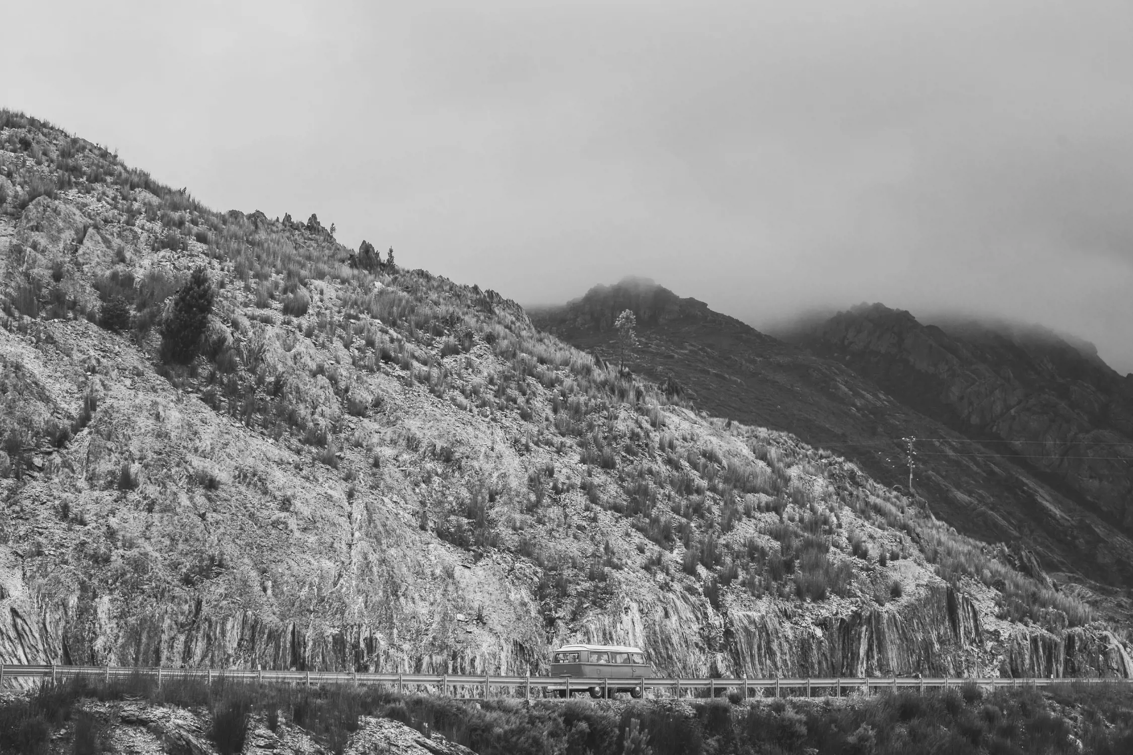 A rocky mountainous landscape with sparse vegetation. A vehicle drives along a winding road carved into the hillside, while mist lingers near the mountain peaks, creating a mysterious and tranquil atmosphere.