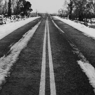 An empty snow-covered road stretches forward in a straight line, flanked by bare trees and a serene, wintry landscape. The white snow contrasts sharply with the dark asphalt and creates a calm, desolate mood.