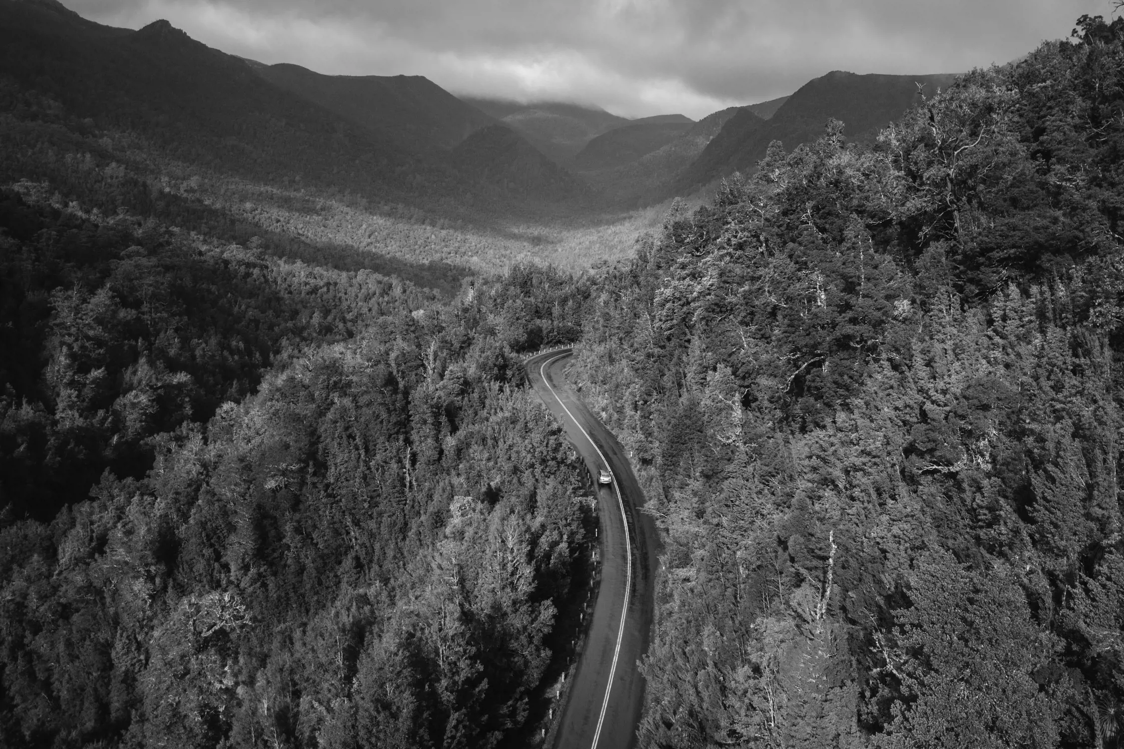 An aerial view of a winding road cutting through a dense forest, with tall trees covering the hilly landscape. Misty mountains rise in the distance under an overcast sky, adding a sense of depth and scale to the scene.