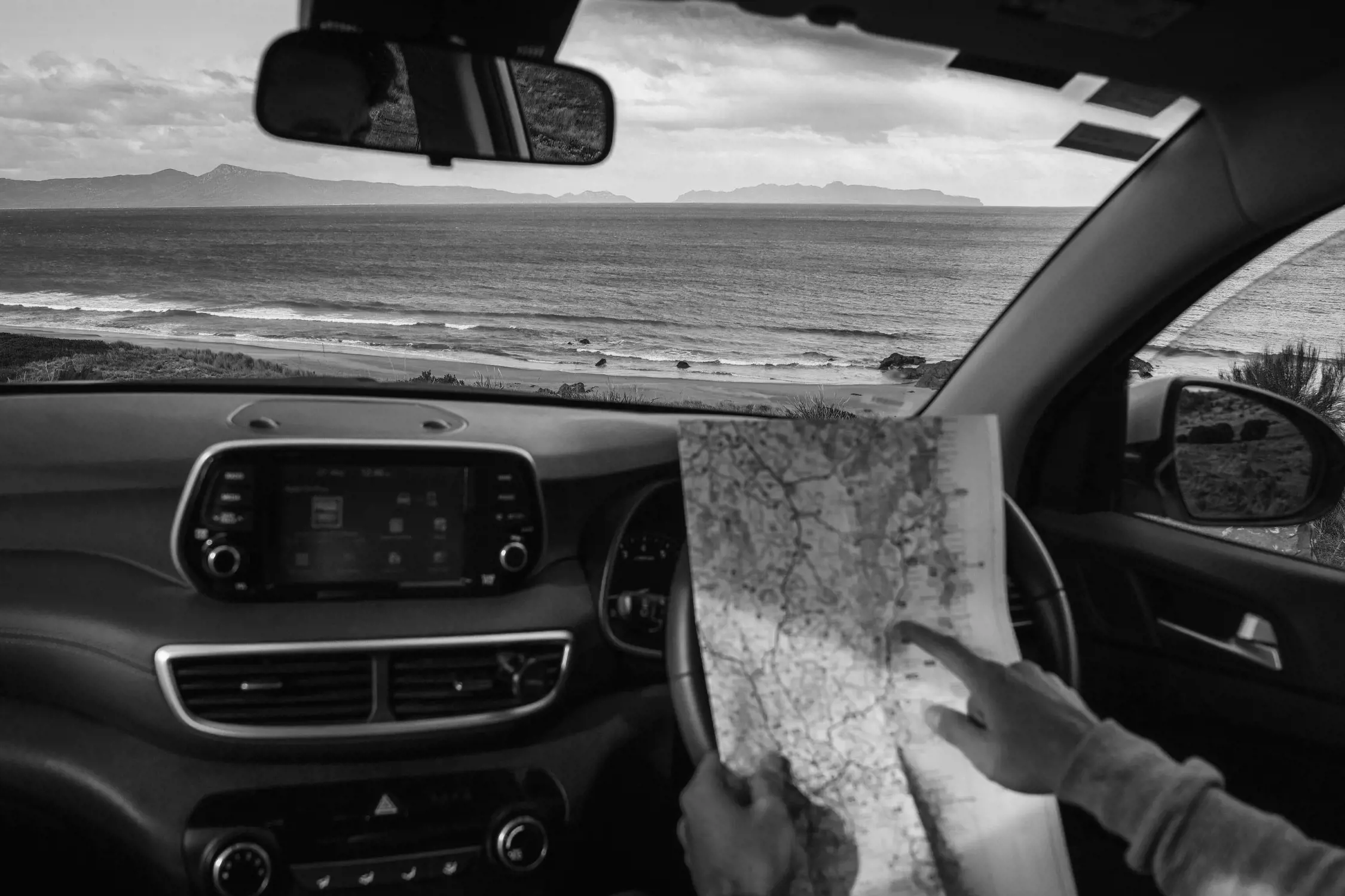 The interior view of a car parked by the beach, with the ocean visible through the windshield. A hand holds a map on the dashboard, suggesting a road trip adventure. The setting is relaxed, with gentle waves and distant islands on the horizon.