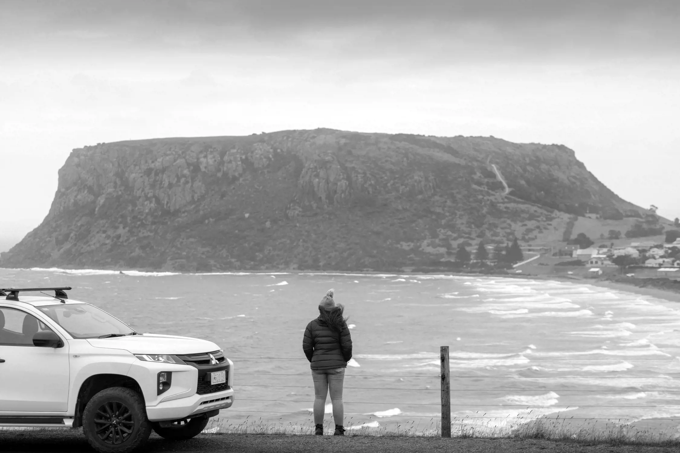 A woman stands on a scenic coastal roadside next to a white SUV. In front of her, the ocean waves crash against the shore, while a prominent, rugged, flat-topped hill, likely The Nut in Stanley, Tasmania, rises in the background. The sky is overcast, adding a serene and dramatic tone to the scene.