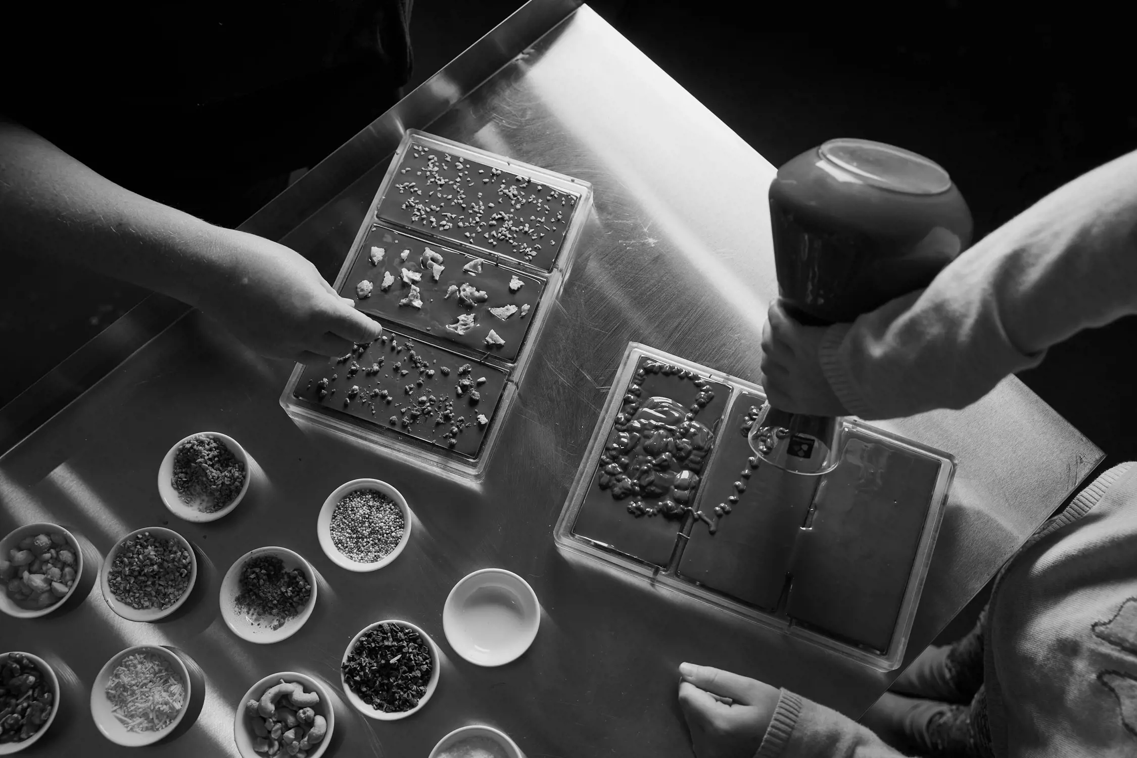 Two people work at a metal table, decorating chocolate bars inside molds. Small bowls filled with a variety of toppings like nuts, seeds, and dried fruits are arranged neatly on the table. One person uses a piping tool to add details to the chocolate.