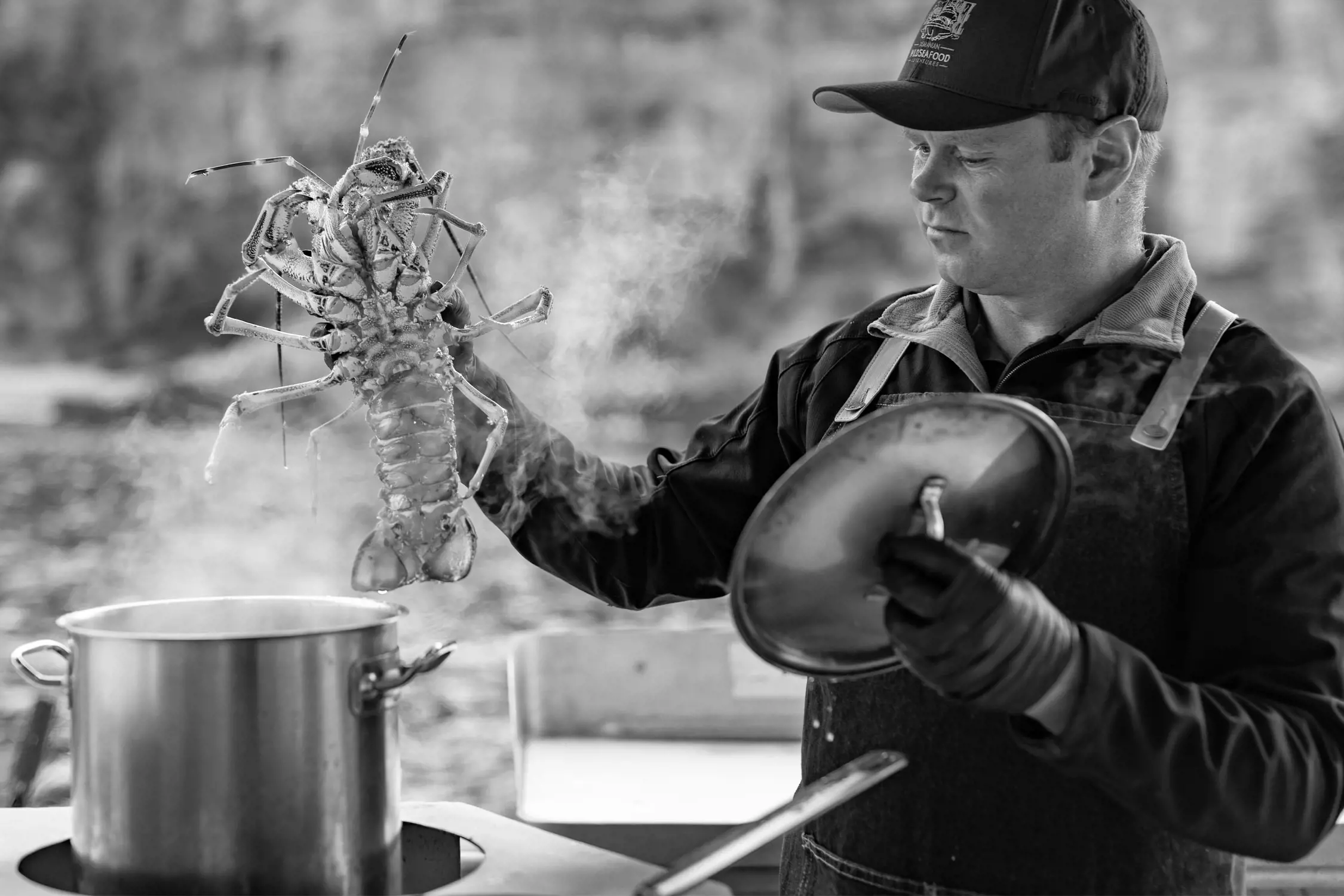 A man wearing an apron and gloves holds a large lobster over a steaming pot of boiling water. He is standing outdoors with a river and rocky shoreline in the blurred background. Steam rises dramatically, creating an atmospheric cooking scene.