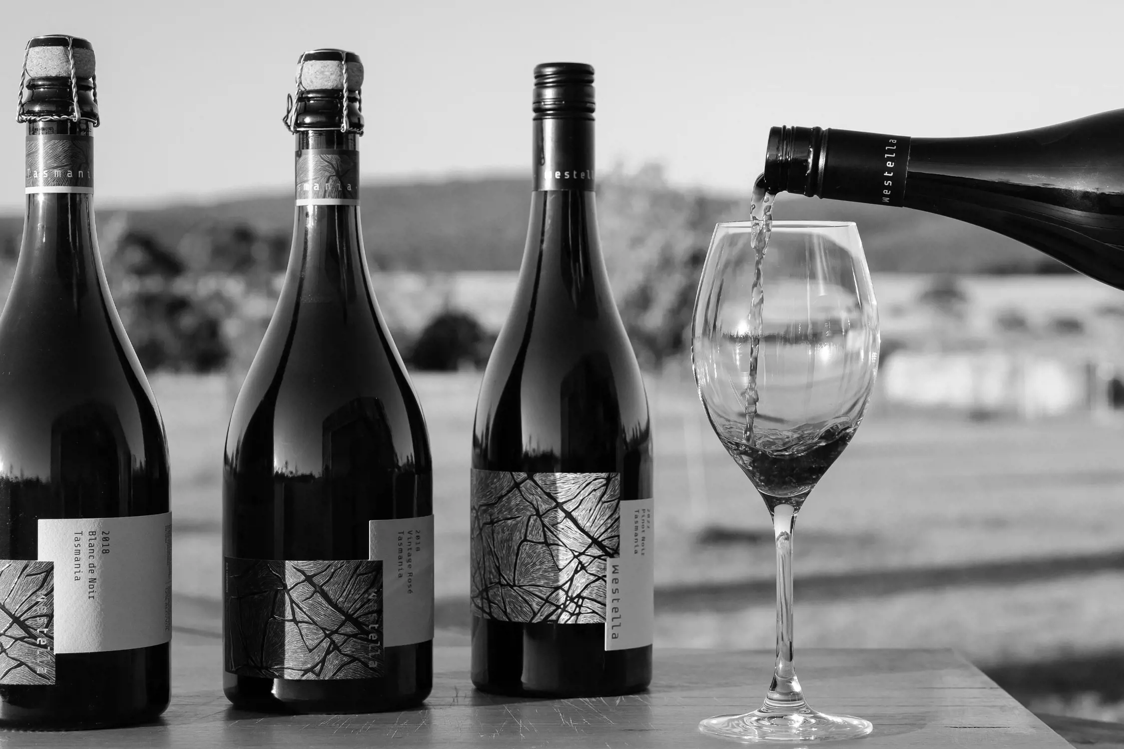 Three elegant bottles of Tasmanian wine are displayed on a wooden table. In the foreground, wine is being poured into a glass, while the background reveals a serene rural landscape with fields and trees, softly blurred for effect.