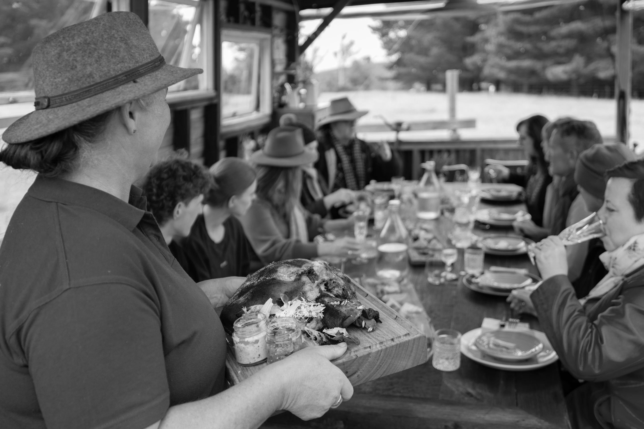 An outdoor dining table under a wooden pavilion is filled with guests enjoying a meal. A woman in the foreground is serving a roasted meat dish on a wooden board. The table is decorated with plates, jars, and glasses, and the diners appear engaged and relaxed in the rural setting.