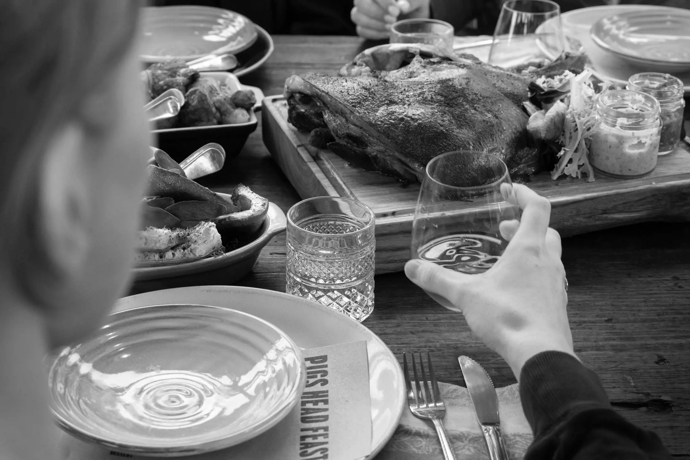 A close-up of a rustic meal at a wooden table. The focus is on a roasted pig’s head presented on a board, accompanied by small jars of condiments. Plates of sides and a hand holding a glass of wine are visible, creating a warm and communal dining atmosphere.