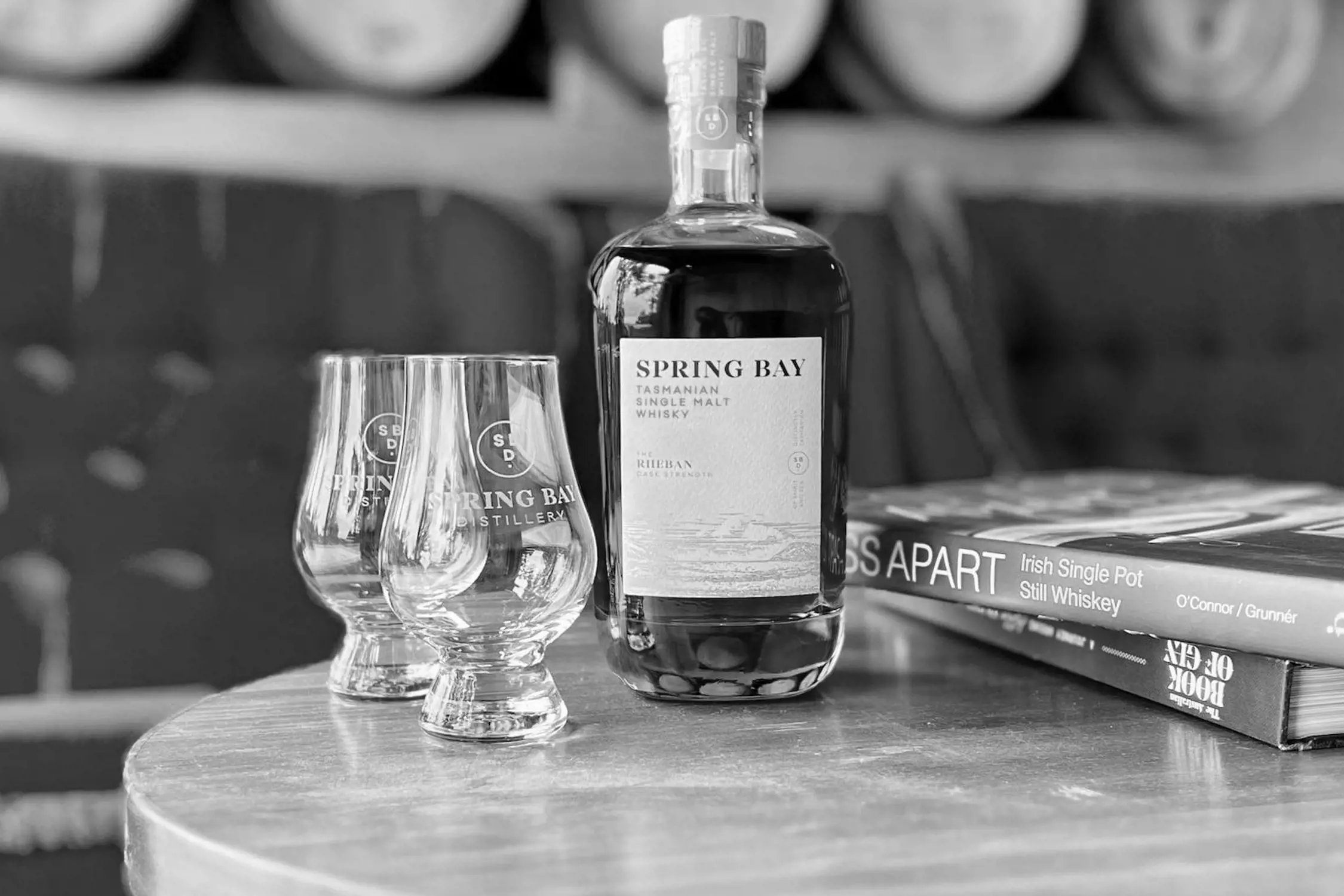 A bottle of Spring Bay Distillery Tasmanian Single Malt Whiskey sits on a round wooden table next to two etched whiskey glasses. Behind the bottle are books on whiskey and blurred barrels, creating a cozy and refined ambiance in the distillery.