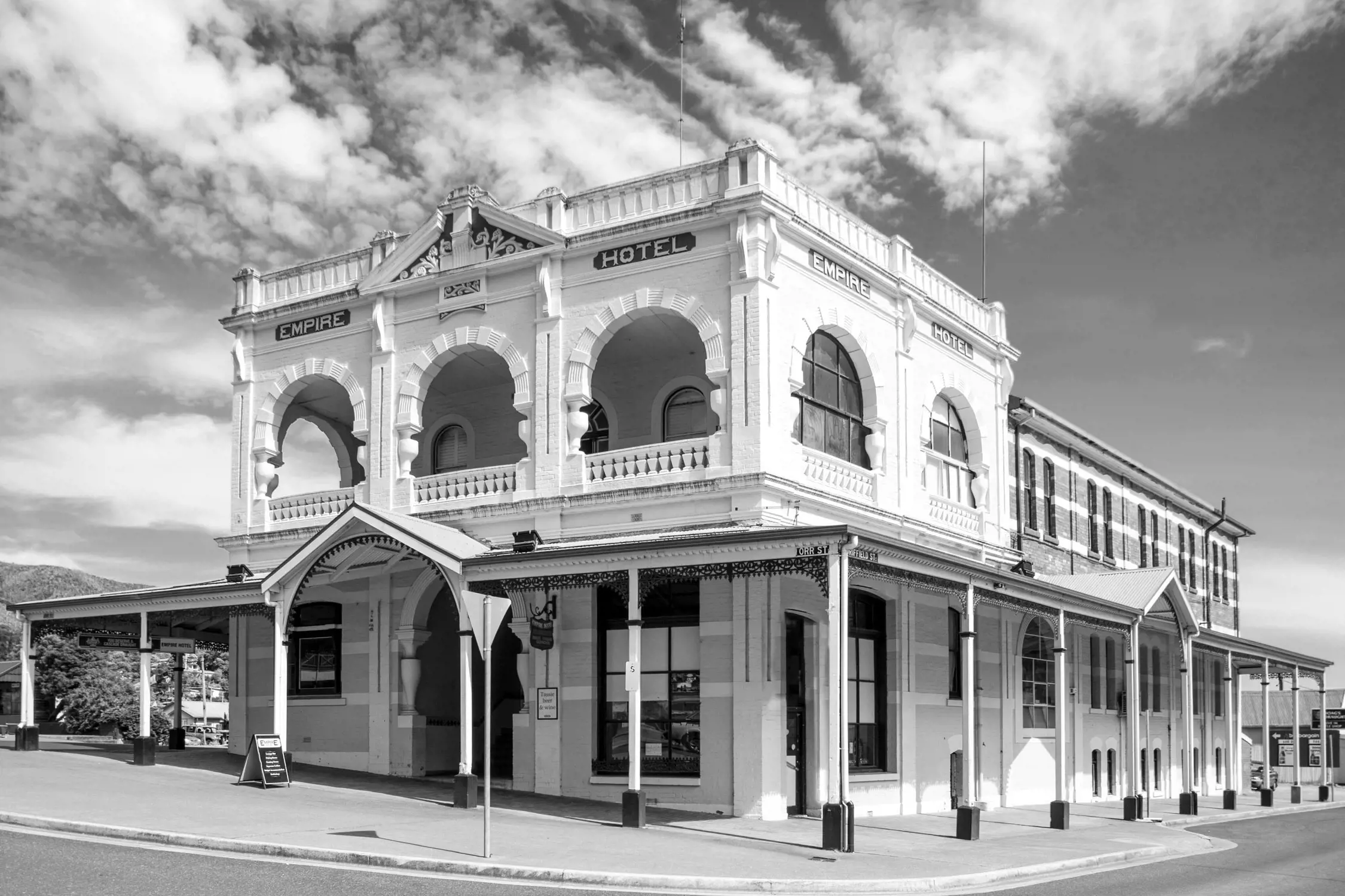 A historic two-story white building labeled 'Empire Hotel' with arched windows and ornate columns. It is situated on a quiet street corner under a partly cloudy sky, reflecting Victorian-era architectural style