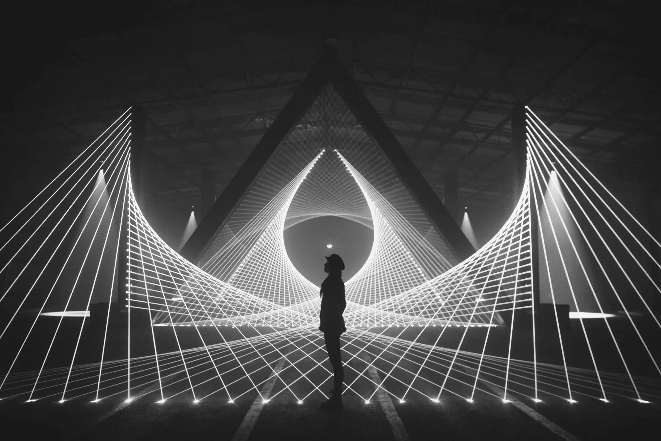 A dark indoor space featuring a geometric light installation of glowing white triangular and circular shapes. A person, silhouetted against the lights, stands in the center, adding a sense of scale to the abstract, immersive design