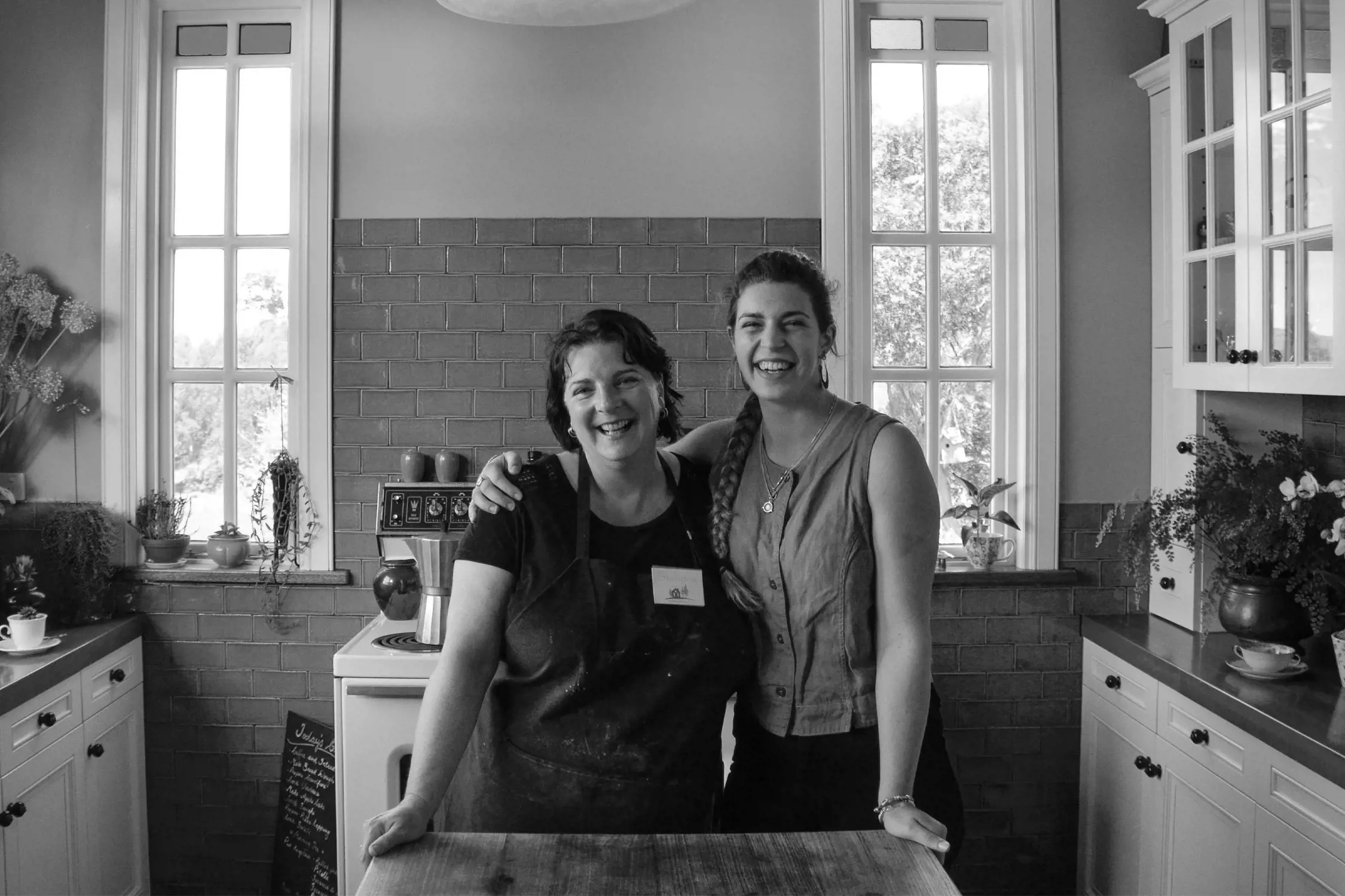 Two women smiling in a cozy farmhouse kitchen with natural light streaming through large windows. One wears an apron, while the other stands beside her, both appearing joyful and welcoming. The kitchen features rustic decor with flowers and wooden countertops