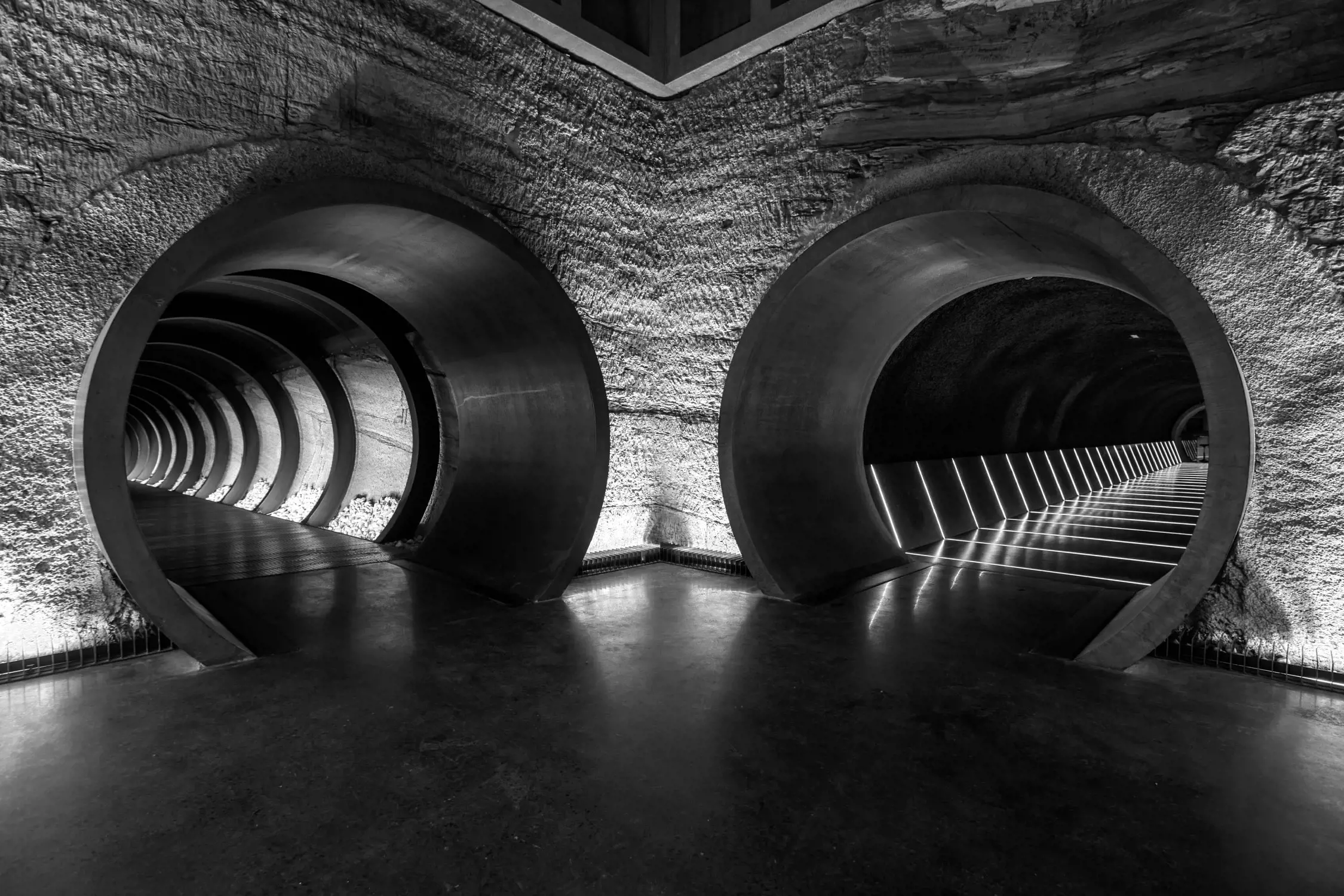 A futuristic underground tunnel with large circular entrances and glowing linear lights along the floor. The lights create a sense of depth, and the textured stone walls add a natural contrast to the sleek, modern design