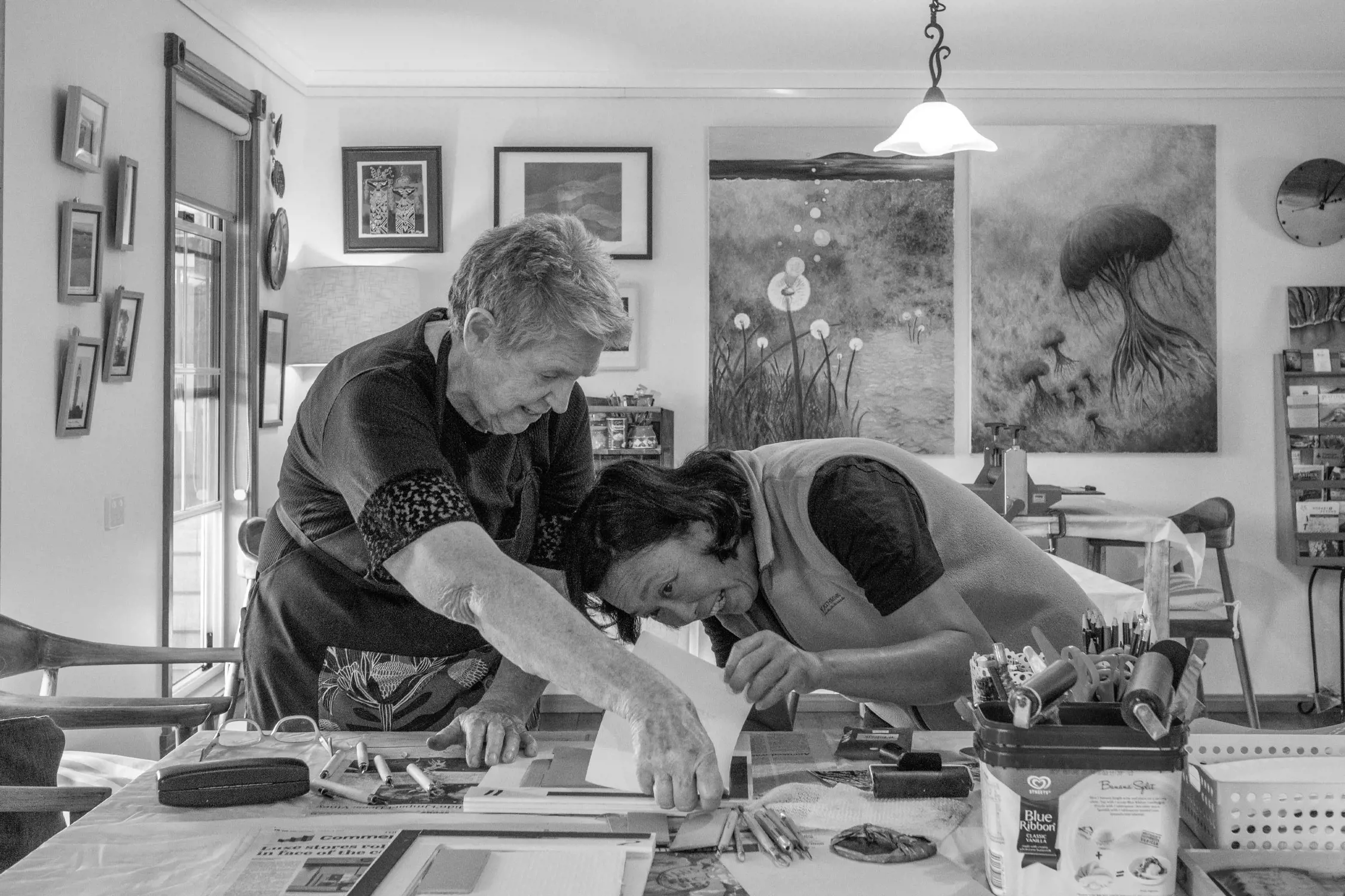 Two women create a linograph in a light filled room tjat has paintings, prints and photos covering the wall. One woman smiles with joy as she lifts the page to revela her print. The table is covered with linograph tools. 