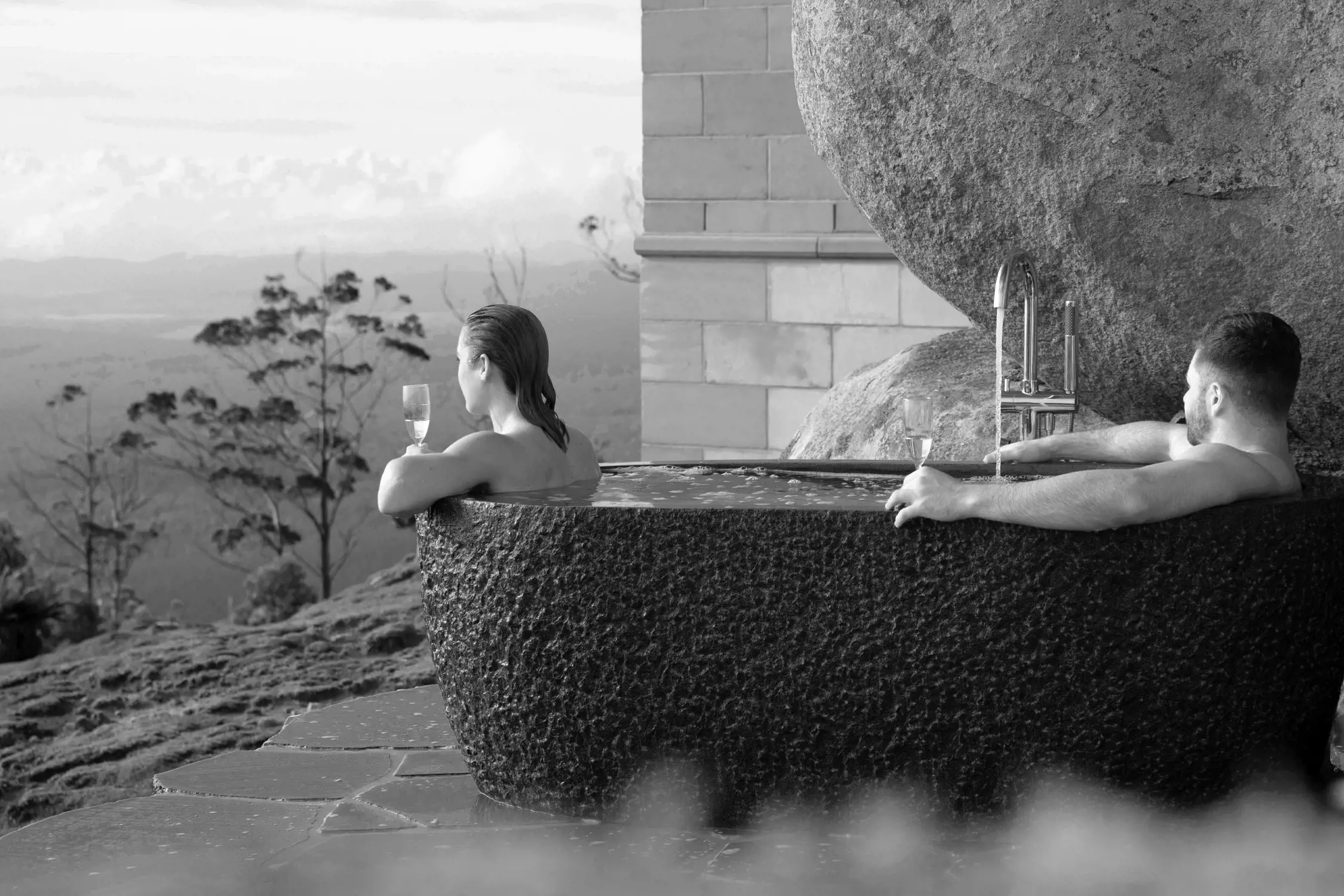 Two people relax in a large, circular outdoor hot tub made from textured stone. Steam rises gently from the warm water, while the pair hold wine glasses and admire a sweeping mountain and valley landscape in the distance. The tub is situated on a rocky hill, adding to the natural, serene atmosphere.