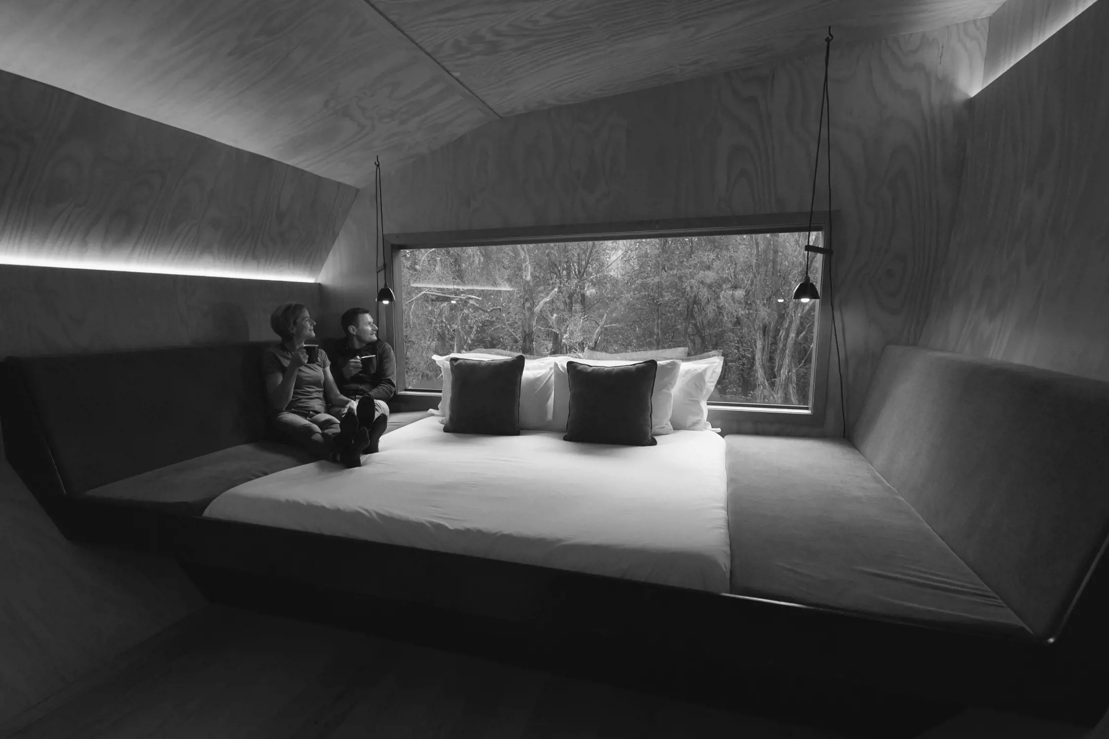 Inside a sleek, minimalist cabin, a large bed with crisp white linens sits against a natural wooden wall. Opposite the bed, a floor-to-ceiling window offers an immersive view of the surrounding dense forest, allowing natural light to fill the room. Two people sit on the bed, enjoying a moment of quiet relaxation