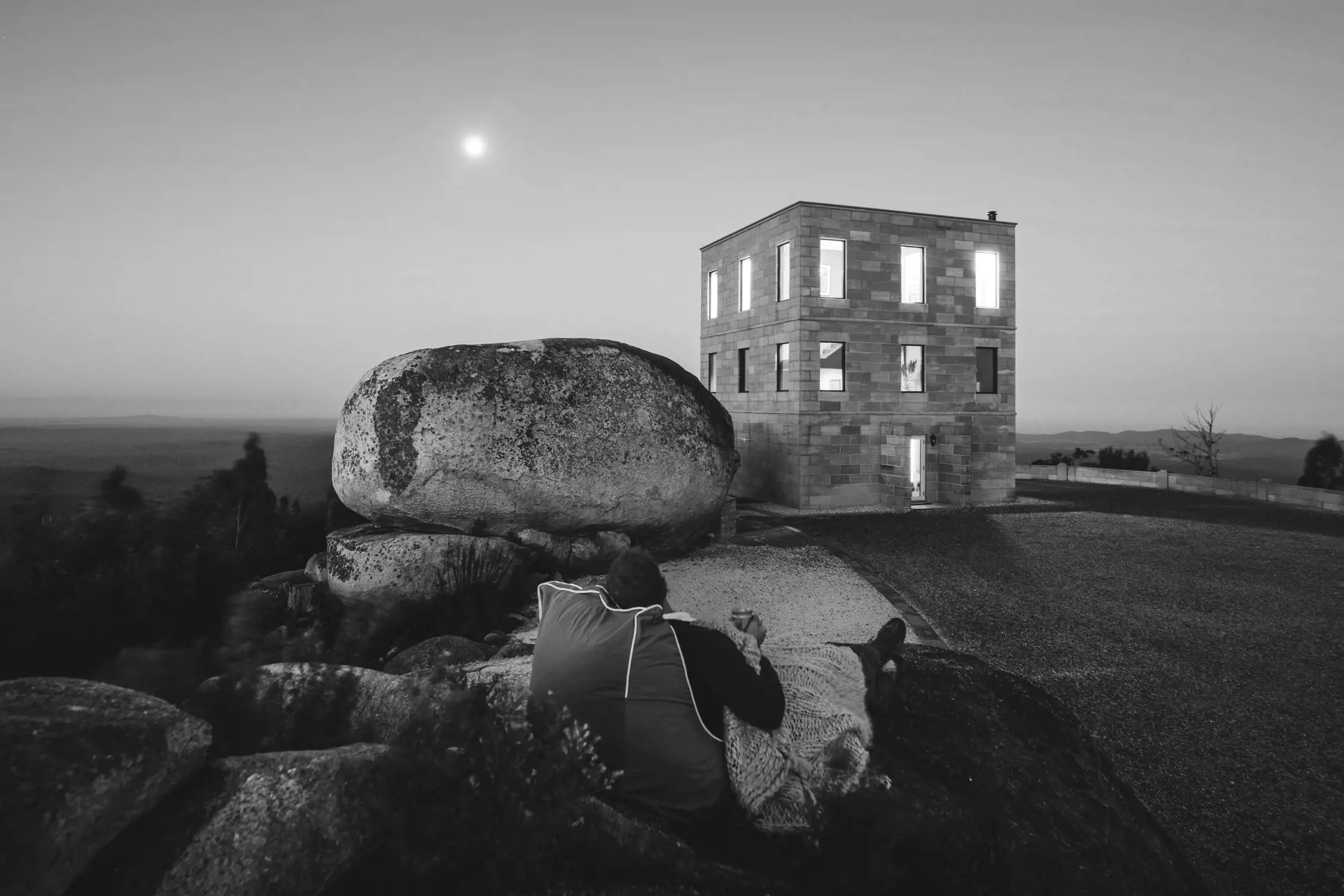 A stone tower with glowing windows stands atop a rocky hillside under a clear night sky. Large boulders surround the structure, adding to its rugged charm. In the foreground, a person wrapped in a blanket leans against a rock, gazing at the illuminated building, which feels isolated yet inviting