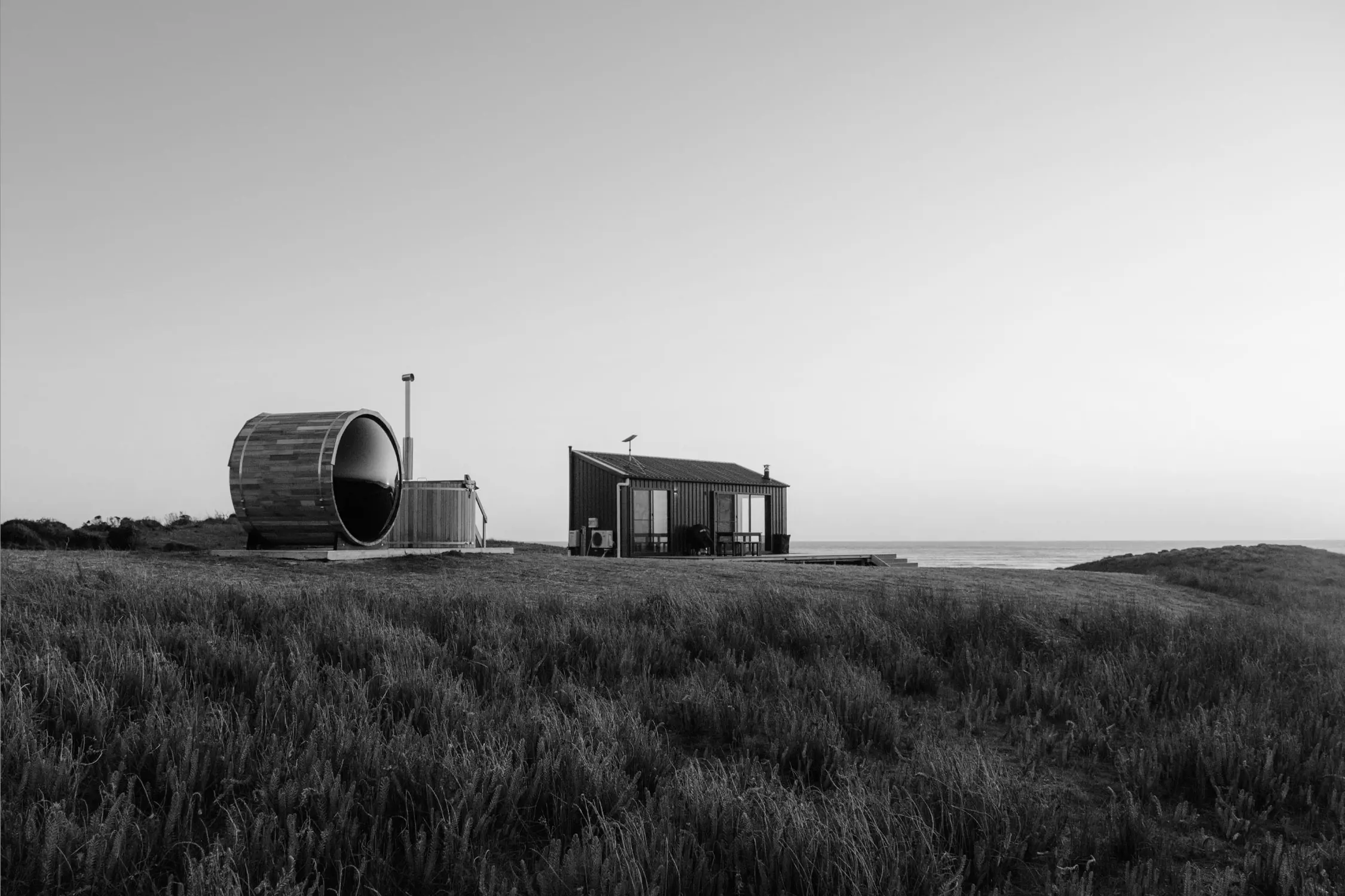 A modern, rustic cabin set in an open coastal field. Next to it is a large, barrel-shaped wooden sauna with a curved roof. The cabin and sauna face the ocean, visible in the distance under a softly lit sky. The landscape is dotted with tall grasses swaying in the wind, creating a sense of peaceful seclusion