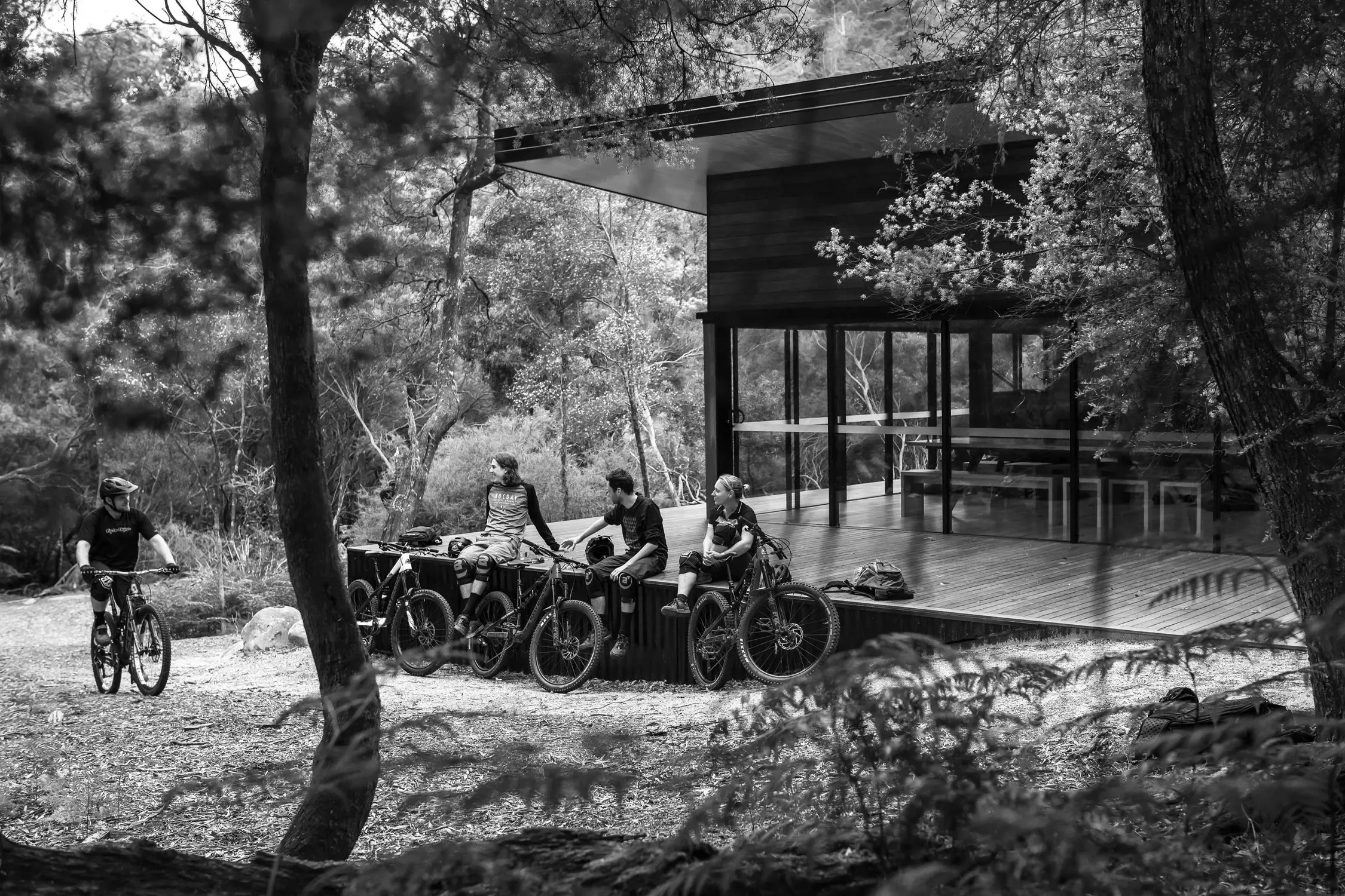 A modern, minimalist outdoor cabin surrounded by dense forest. A group of cyclists rests nearby, with some sitting on their bikes and one actively riding. The cabin has large glass windows that reflect the surrounding trees, blending seamlessly with the natural environment