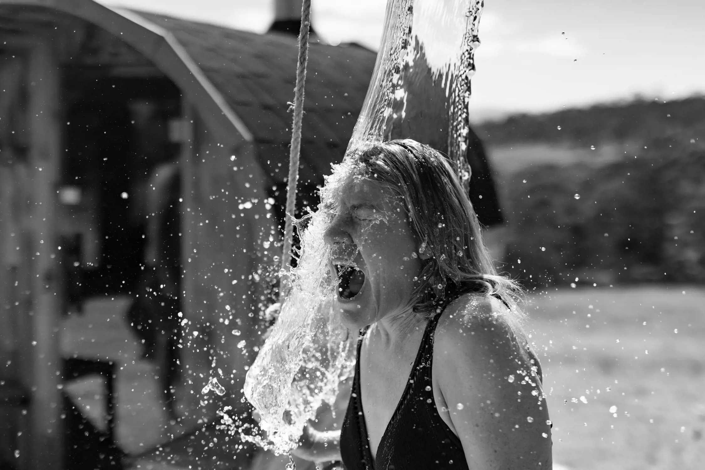 A woman is captured under a cold outdoor bucket shower, water splashing dramatically as it cascades over her head and face. She wears a swimsuit and appears to be gasping or shouting, vividly expressing the shock of the cold water. A small circular structre of a sauna with a chimney is behind her.